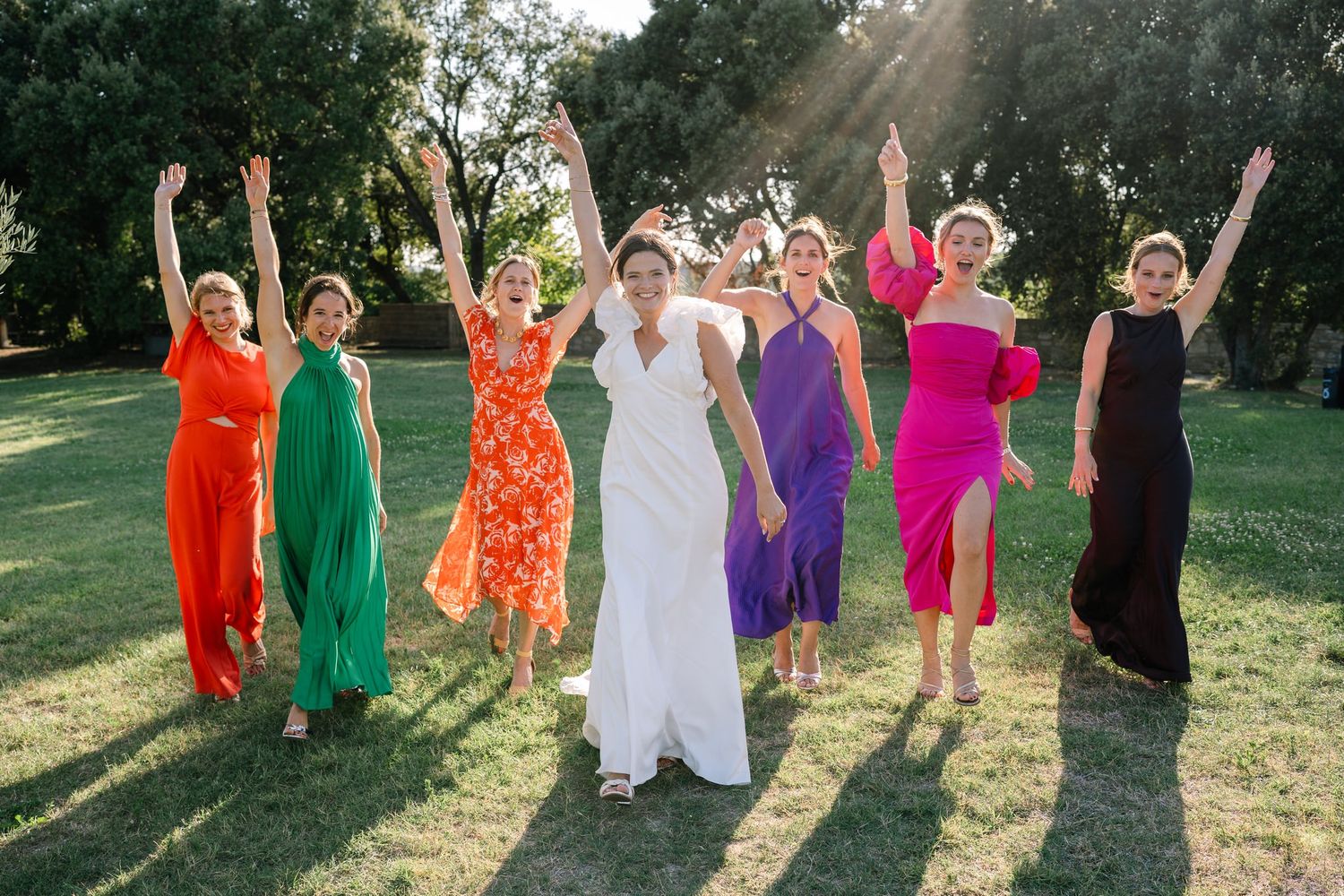 Bridesmaids in colorful dresses jumping joyfully on a sunny lawn.