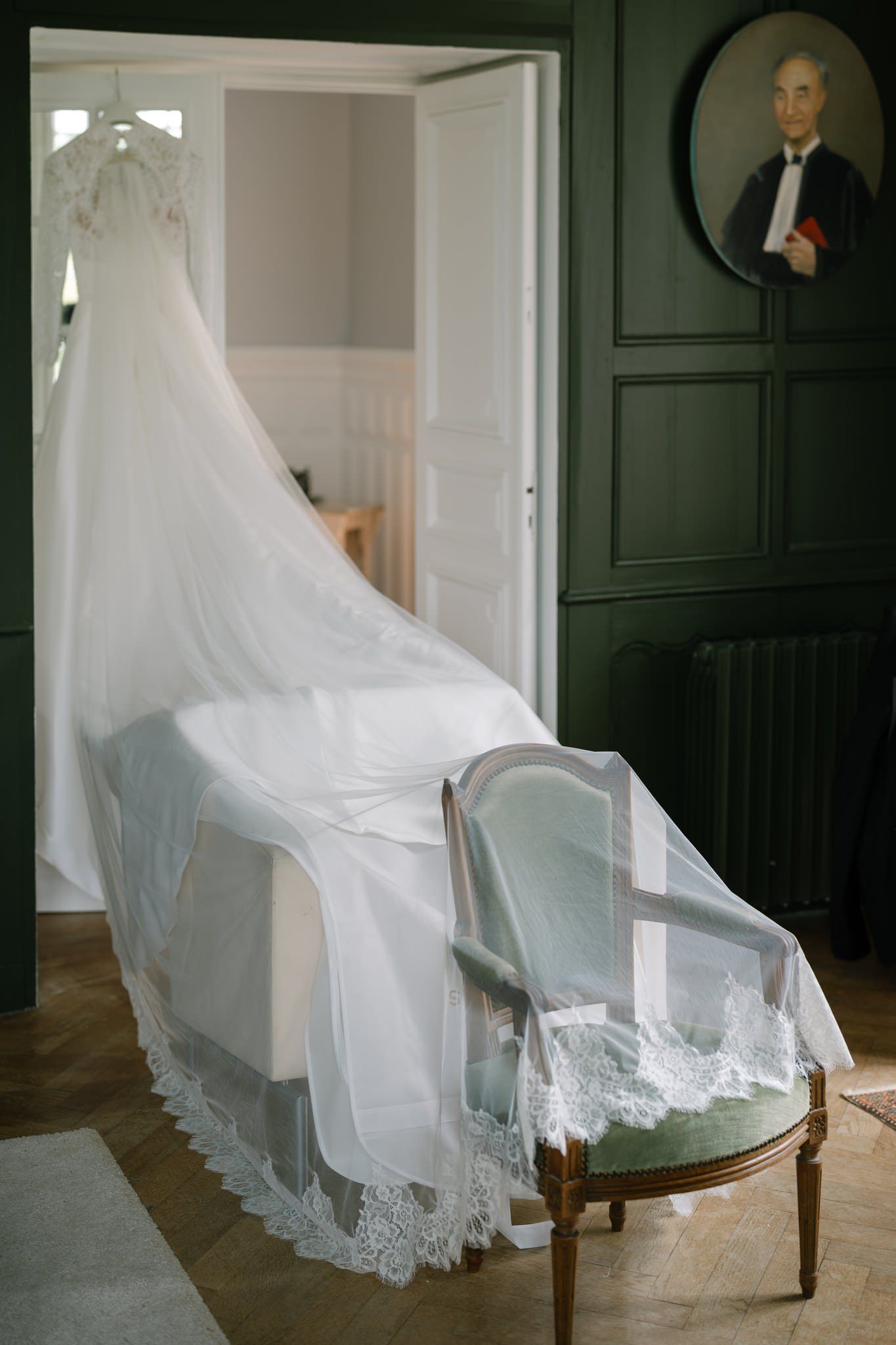 Flowing white wedding veil draped over antique blue chair against dark wall.