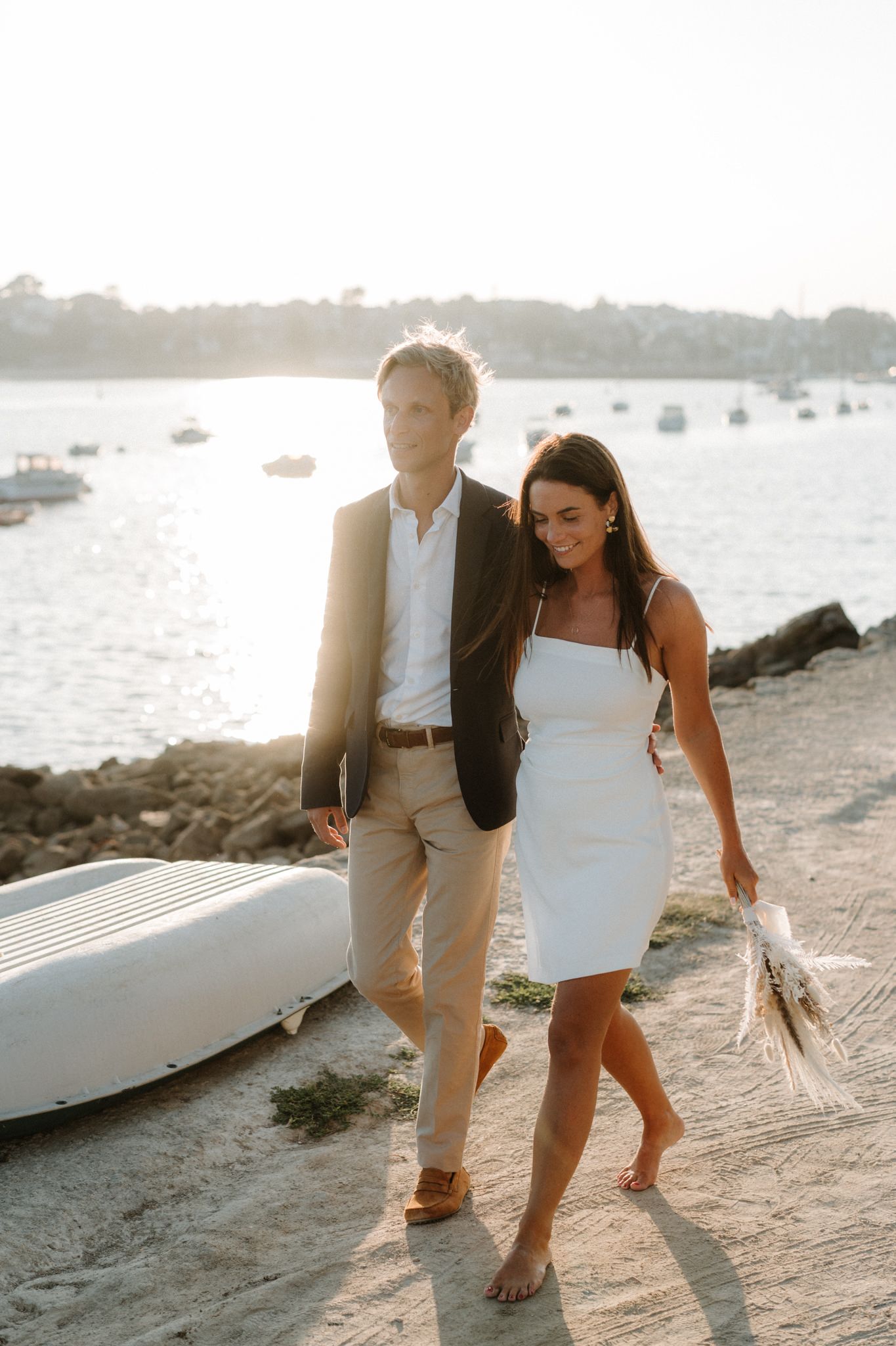 A couple walking barefoot along a beach at sunset with boats in the harbor behind them.