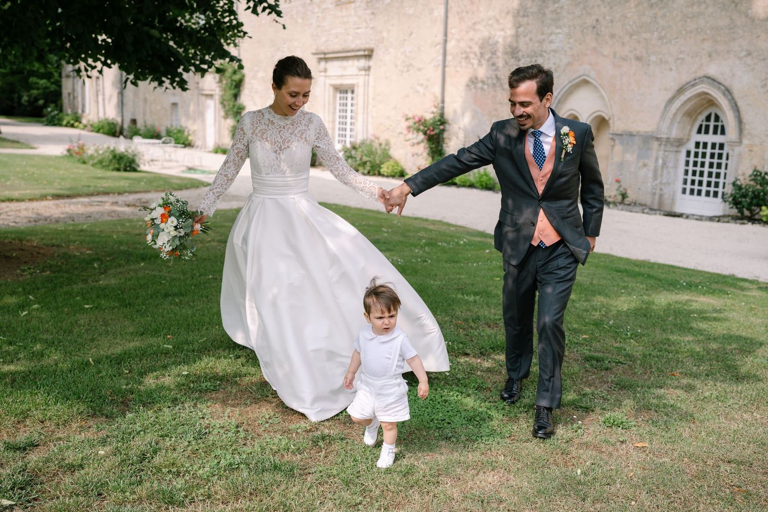 Wedding party walks across a green lawn in front of historic stone buildings.