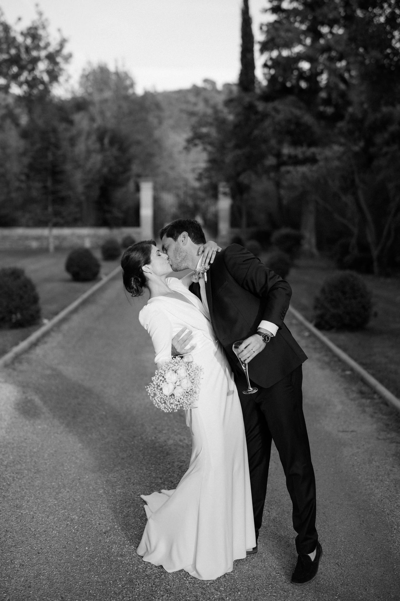 Elegant black and white photo of couple sharing champagne kiss on garden path.