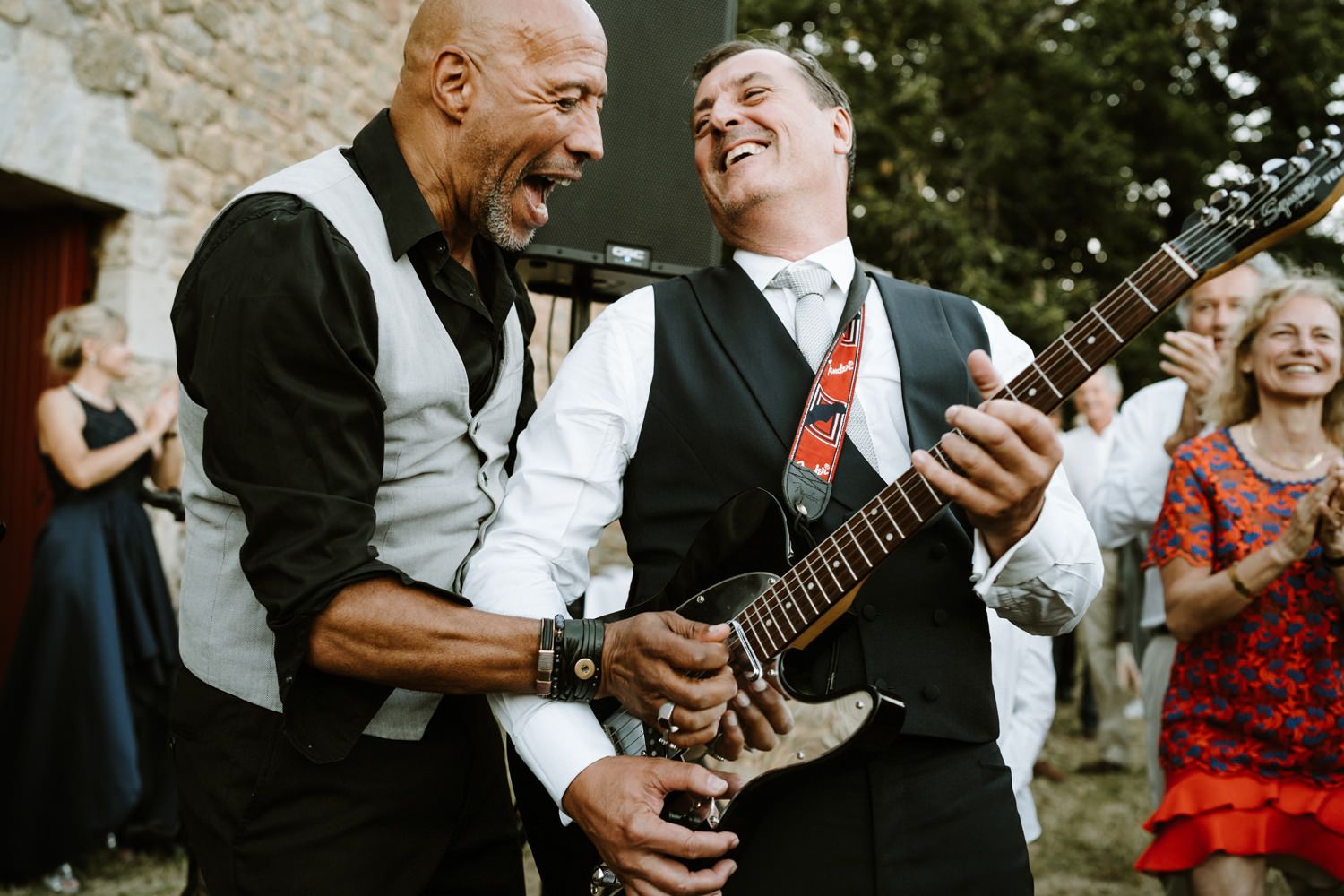 Musicians performing with guitars at outdoor wedding reception near stone wall.