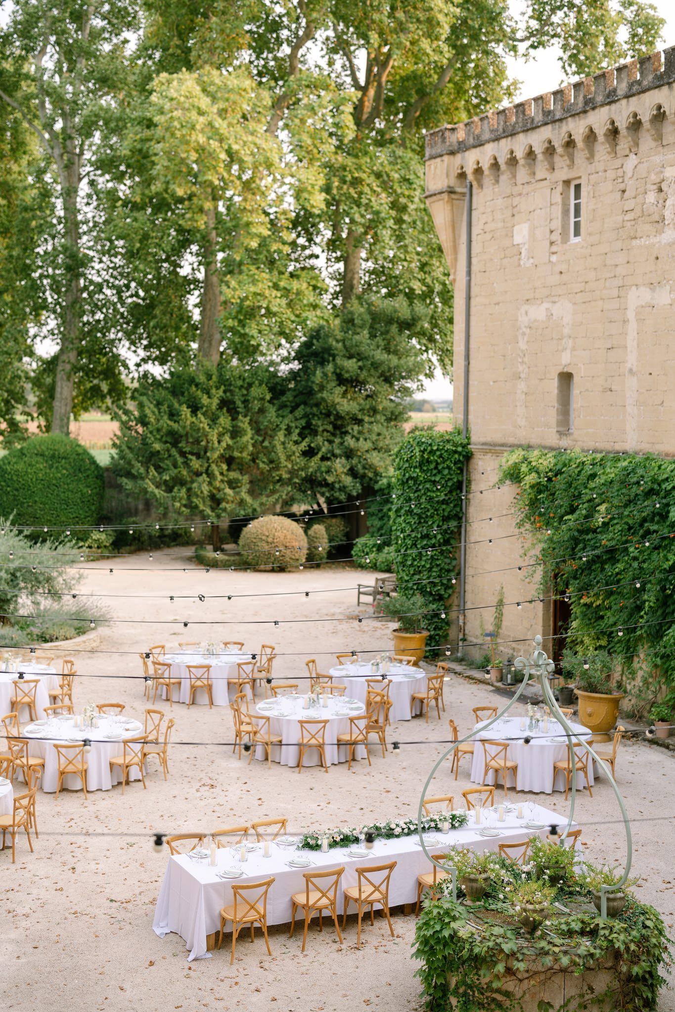 Outdoor wedding reception setup with round tables and wooden chairs against a historic stone building.