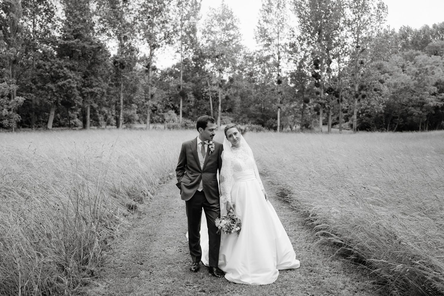 Wedding couple walks through tall grass in a serene countryside setting.