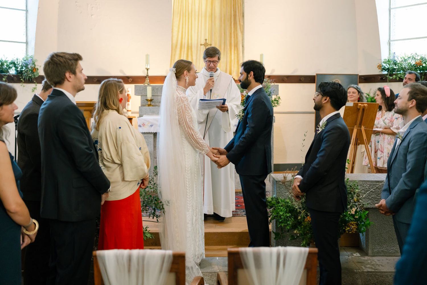 Wedding guests gather in a bright church interior with white walls and natural lighting.