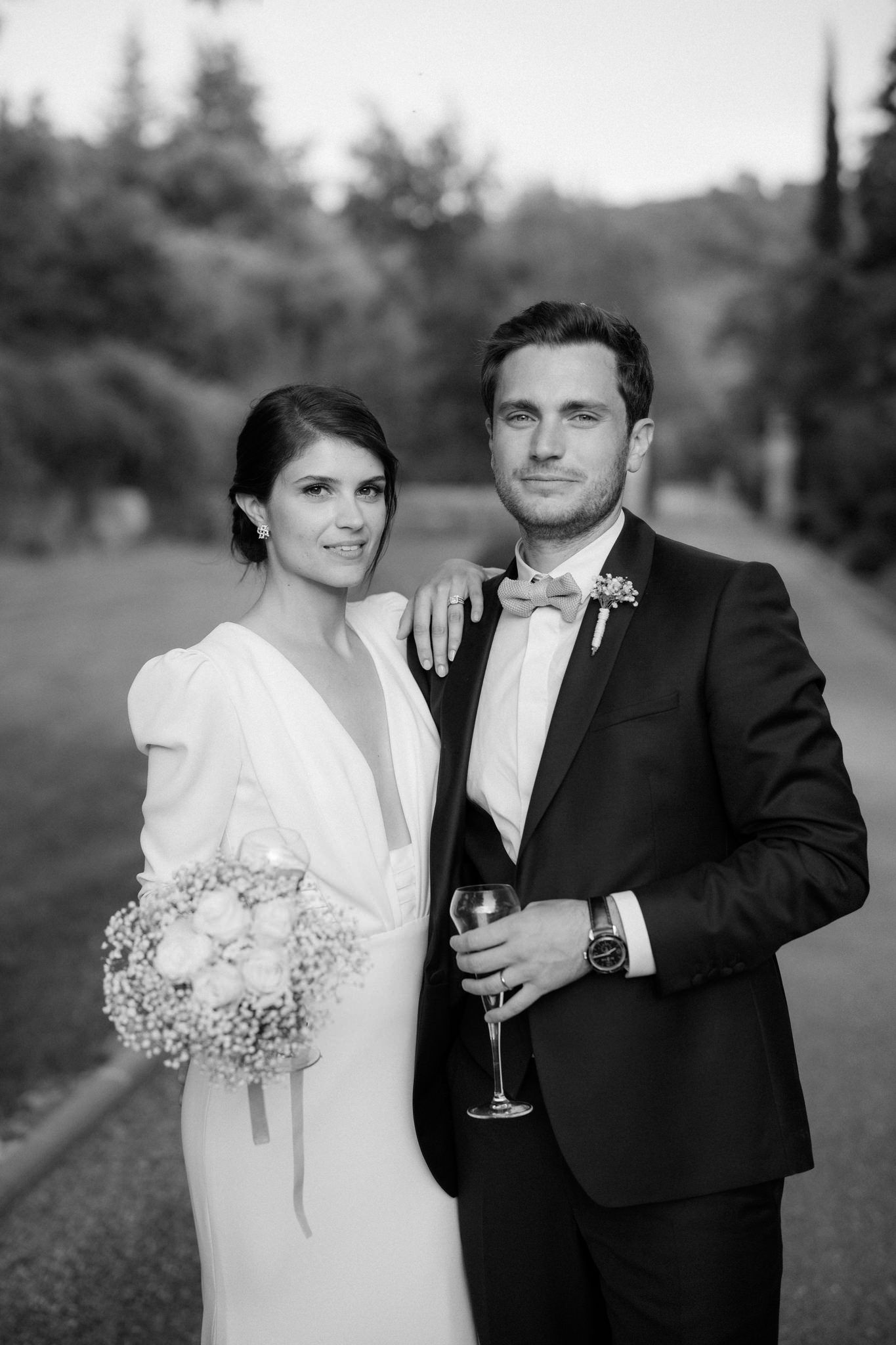 Black and white wedding portrait of a couple holding bouquets and champagne.