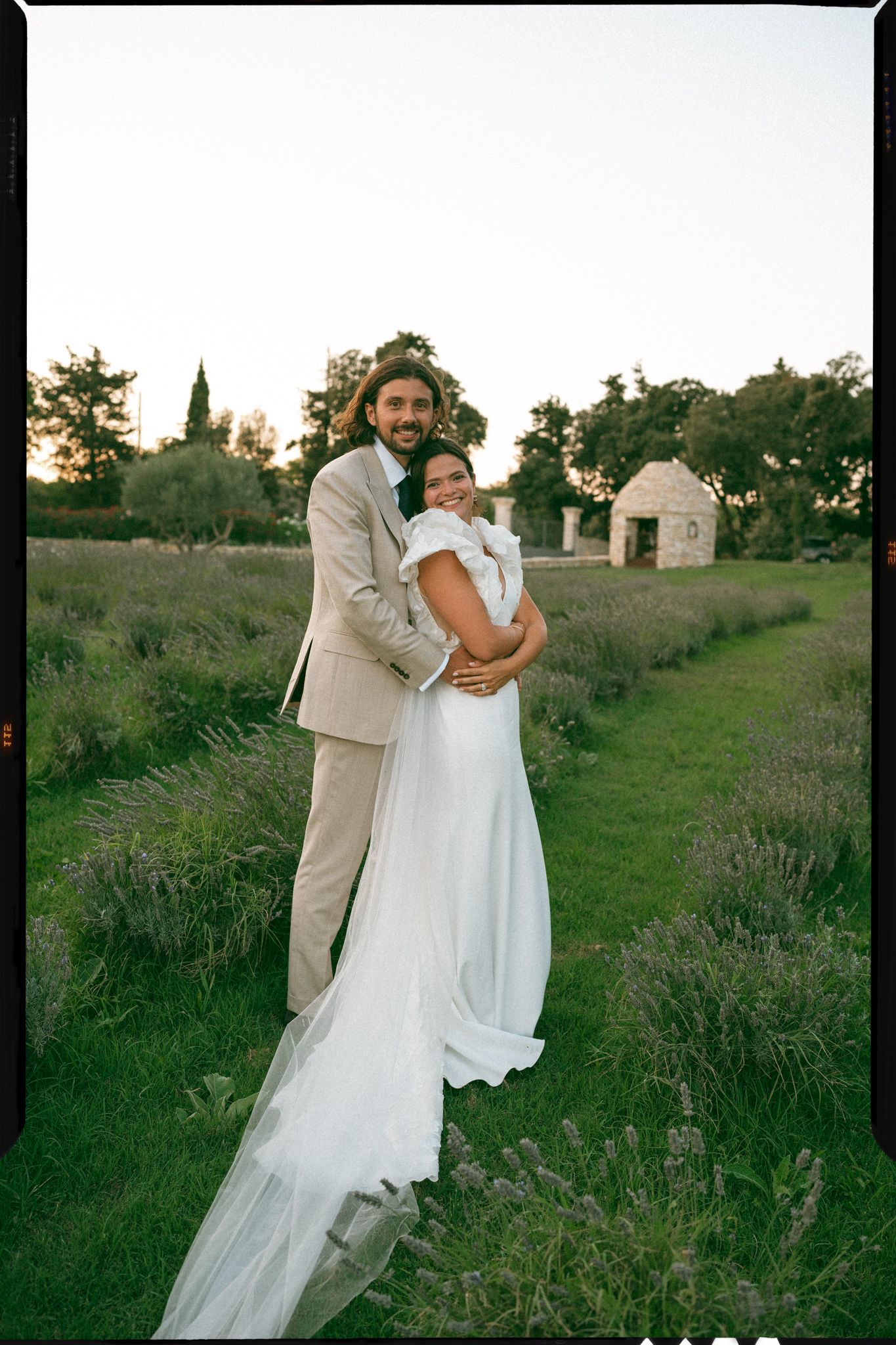 A couple embraces in a romantic outdoor wedding setting with a rustic stone building and greenery in the background at sunset.