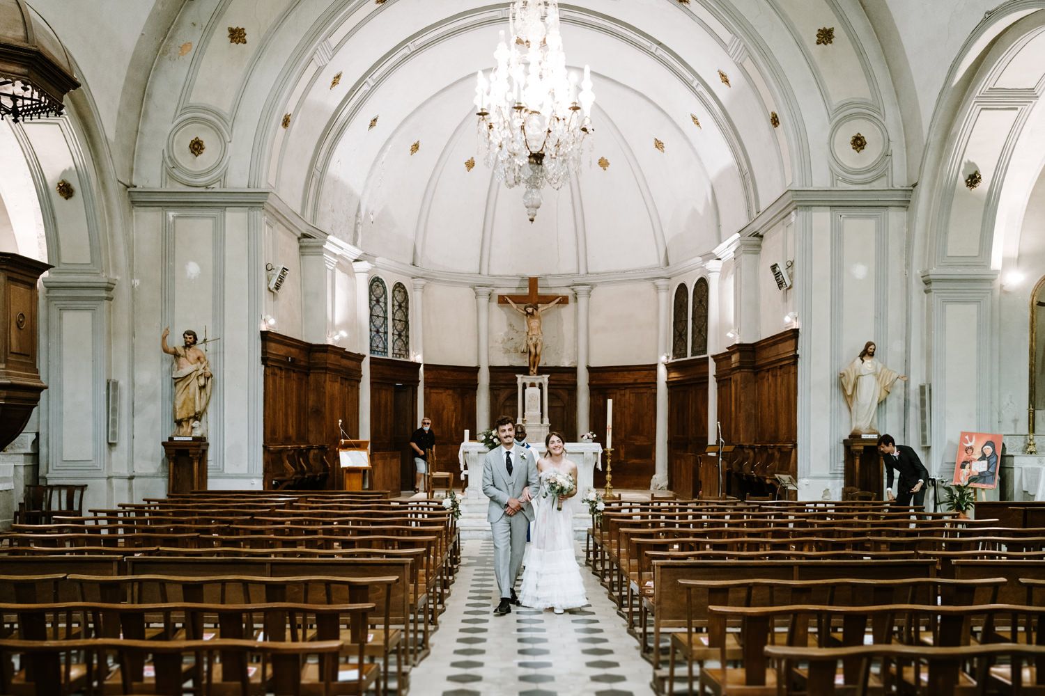 A couple walking down the aisle of an ornate church with vaulted ceilings and chandelier.