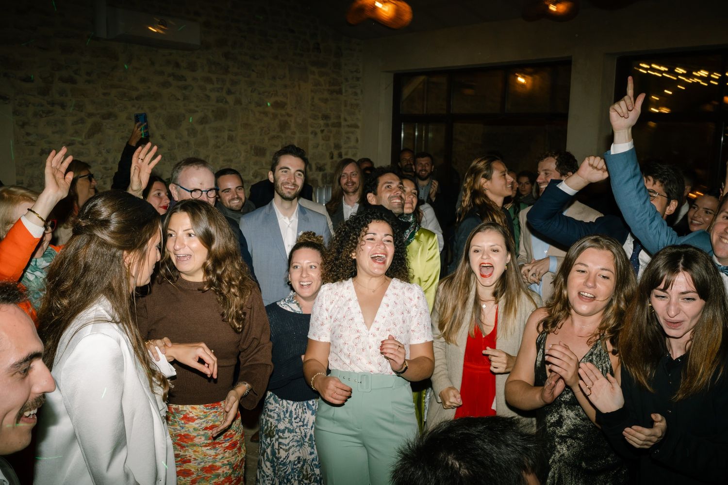 Large group of wedding guests dancing and celebrating together under indoor lighting.