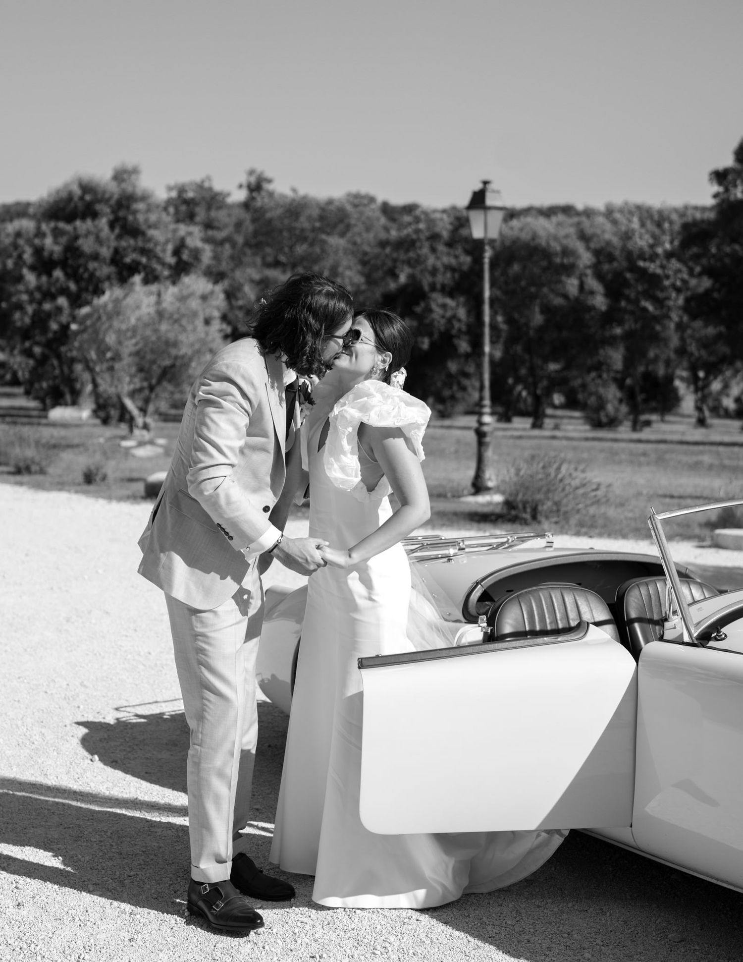 Wedding couple in white attire posing next to a classic white convertible car.
