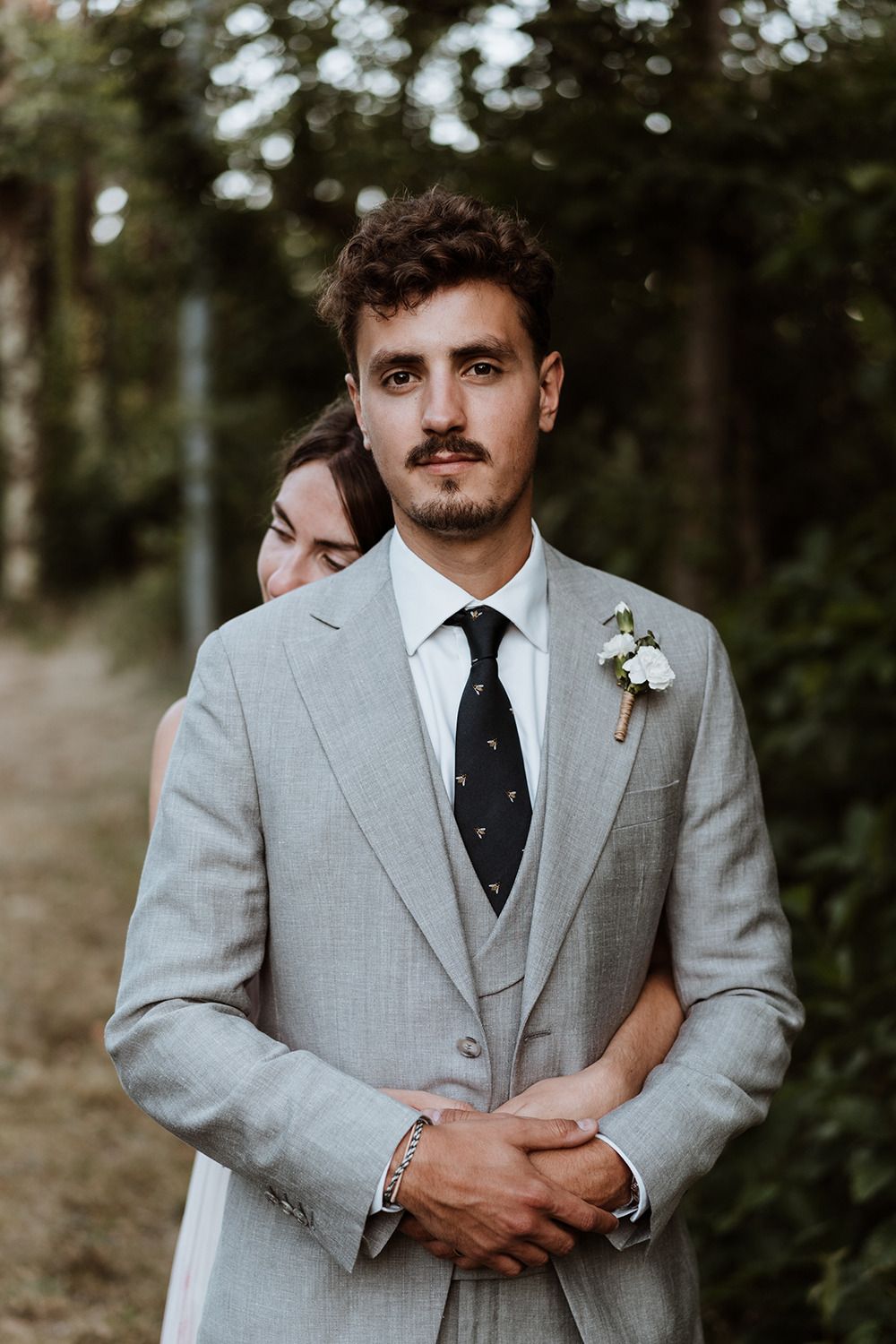 A groom in a light grey three-piece suit with a black tie poses in a natural outdoor setting.