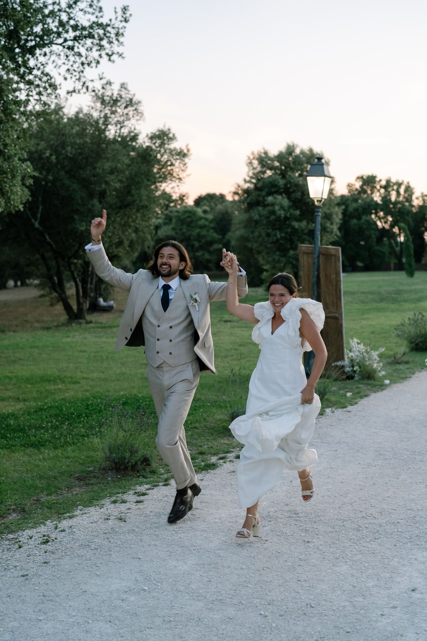 Newlyweds jump playfully together on a white gravel path surrounded by trees.