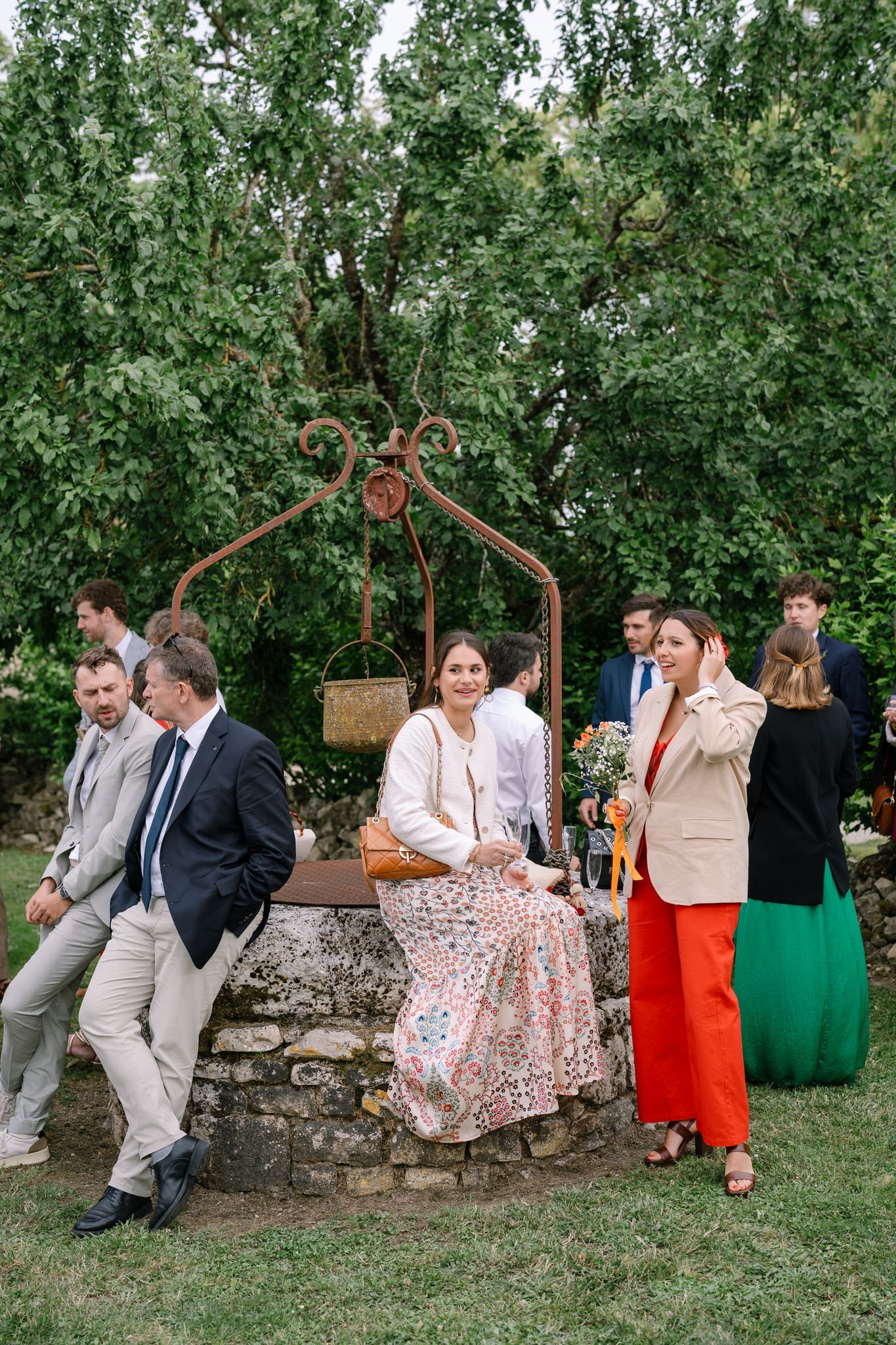 Wedding guests in colorful dresses and suits relaxing on stone wall in garden setting.