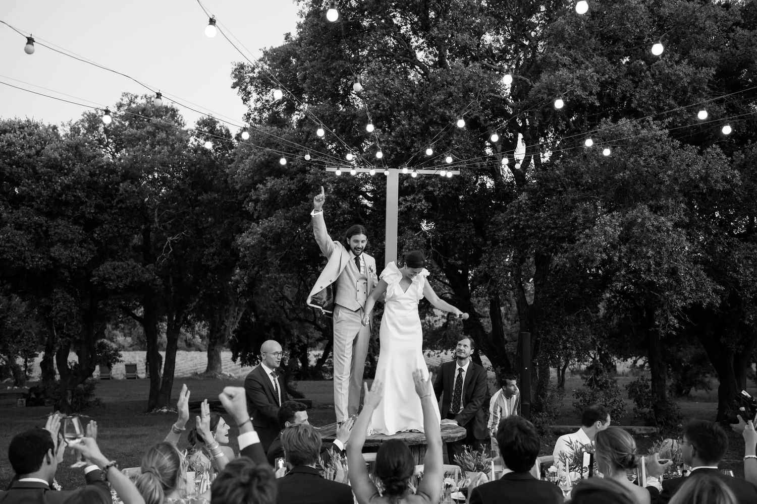 Black and white photo of wedding guests celebrating under string lights at night.