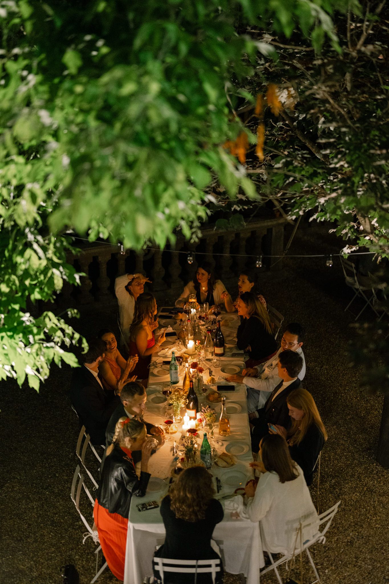 Evening outdoor wedding dinner under trees with candlelit table settings.