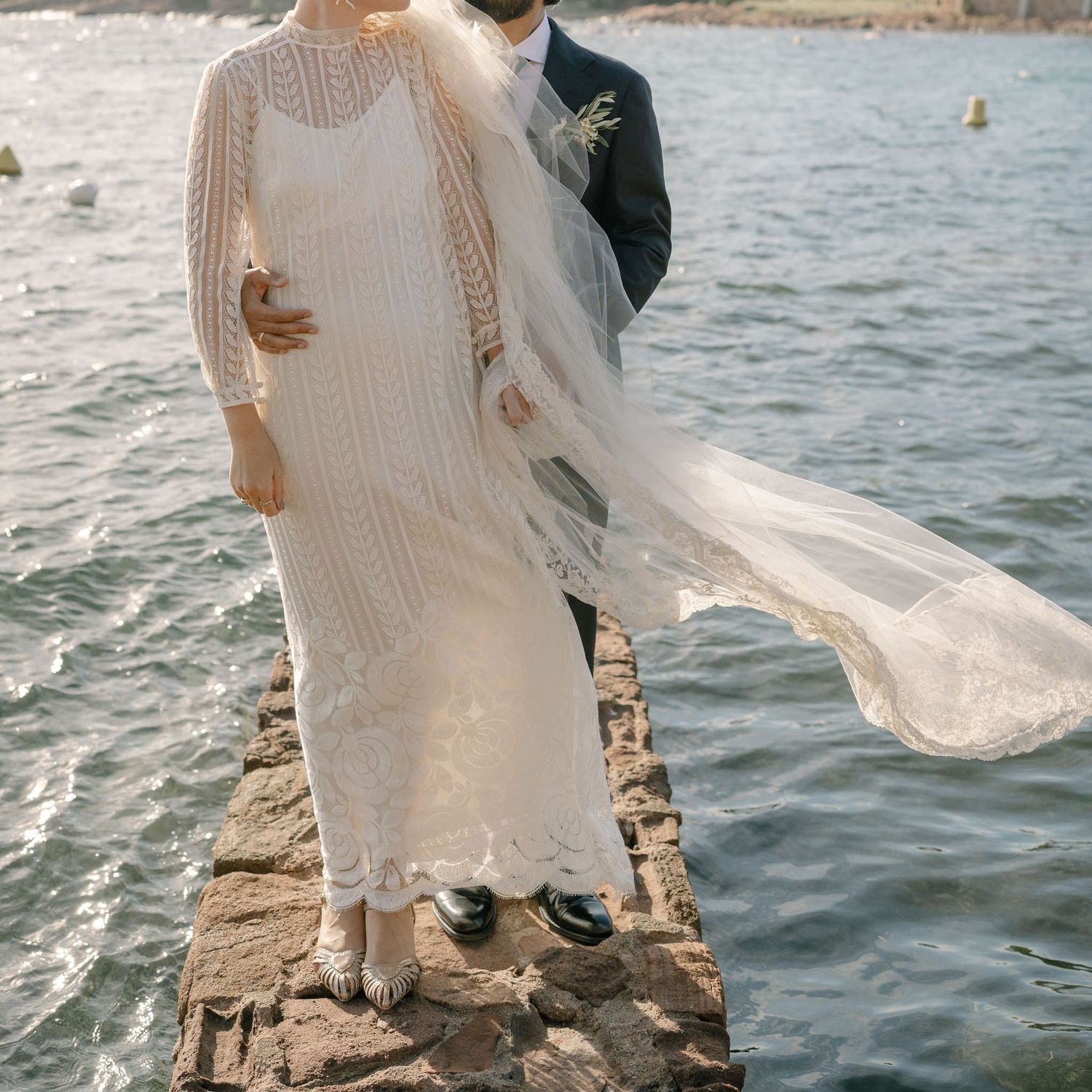 A wedding couple stands on coastal rocks with a flowing veil against the blue Mediterranean Sea.