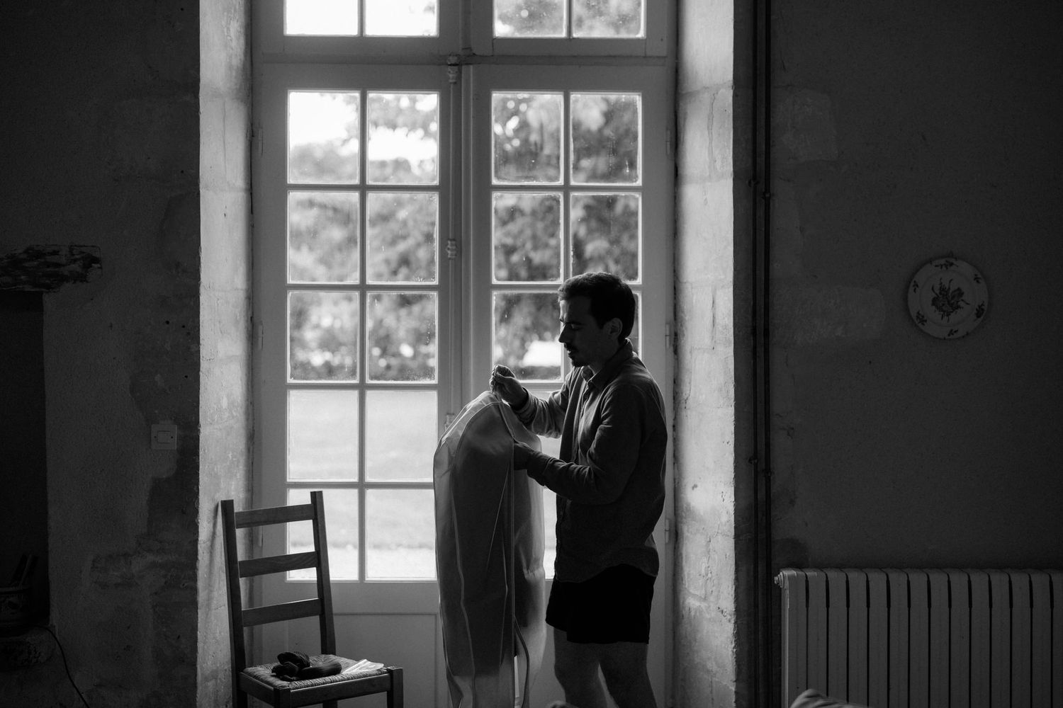 Bright window light streams into an empty room with dark walls and wooden chair.