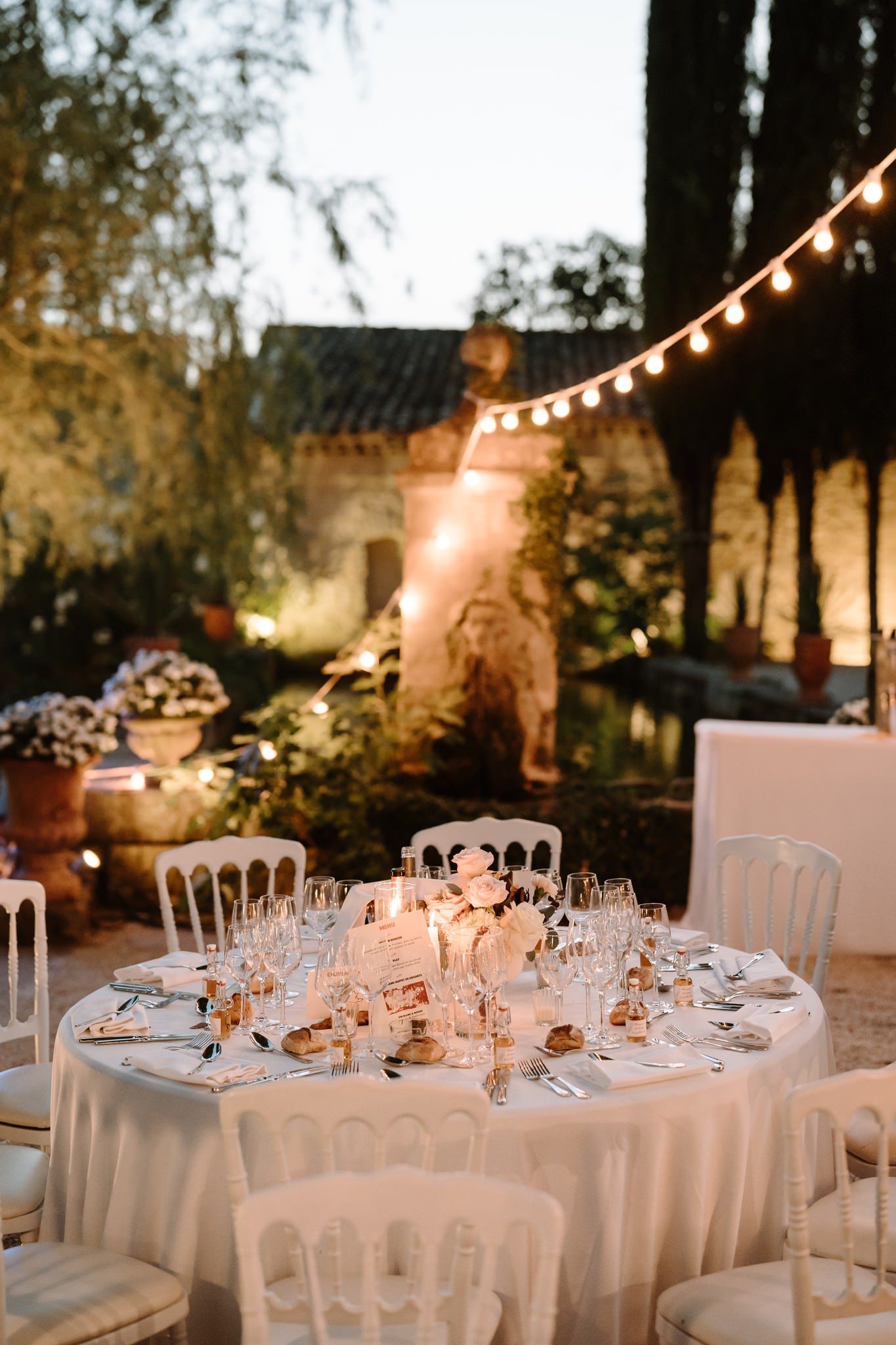 Elegant outdoor wedding reception table set with candles, glassware, and floral centerpieces under string lights at dusk.