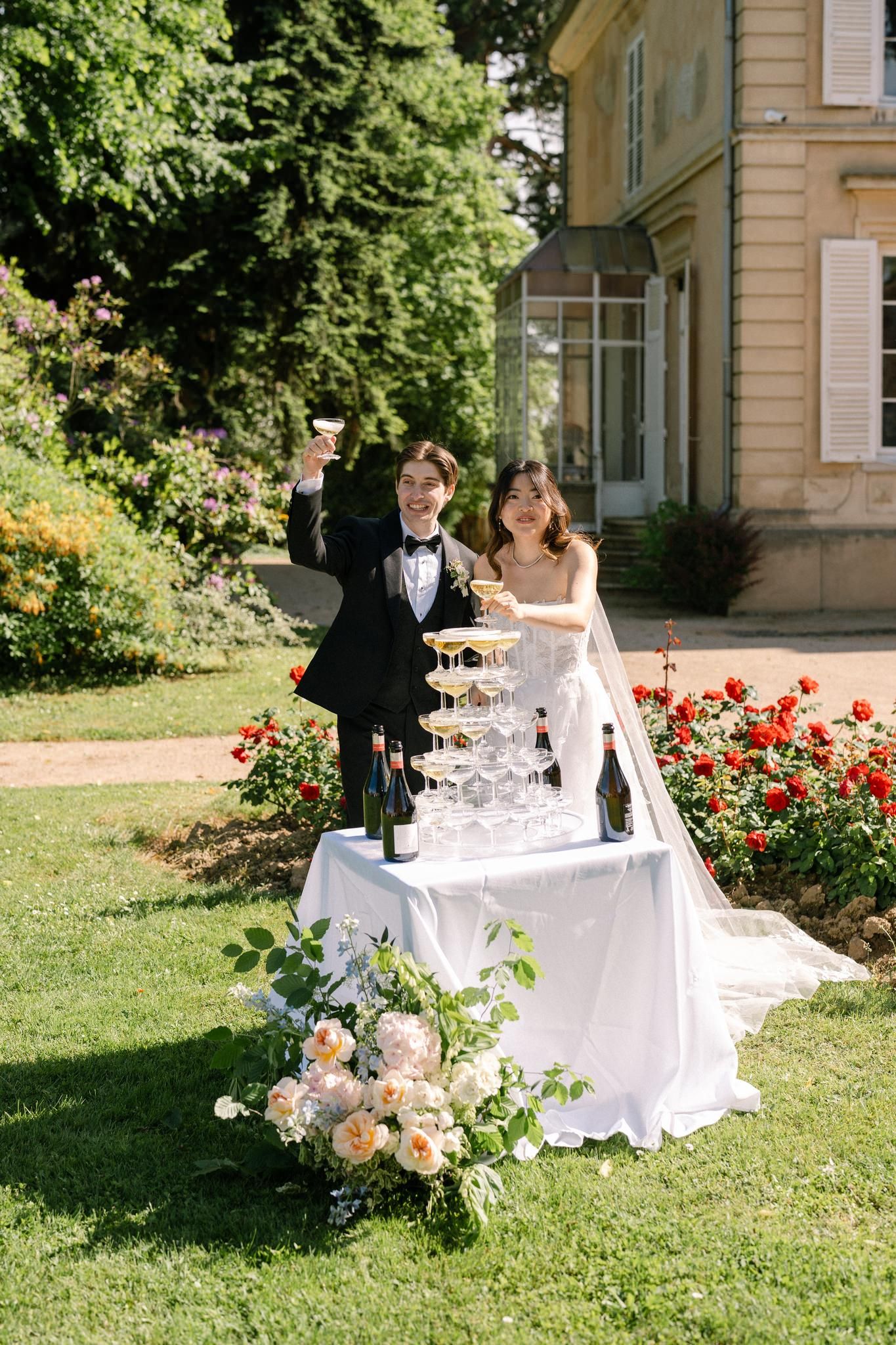 Bride and groom toasting with champagne tower at outdoor wedding reception in elegant garden setting.