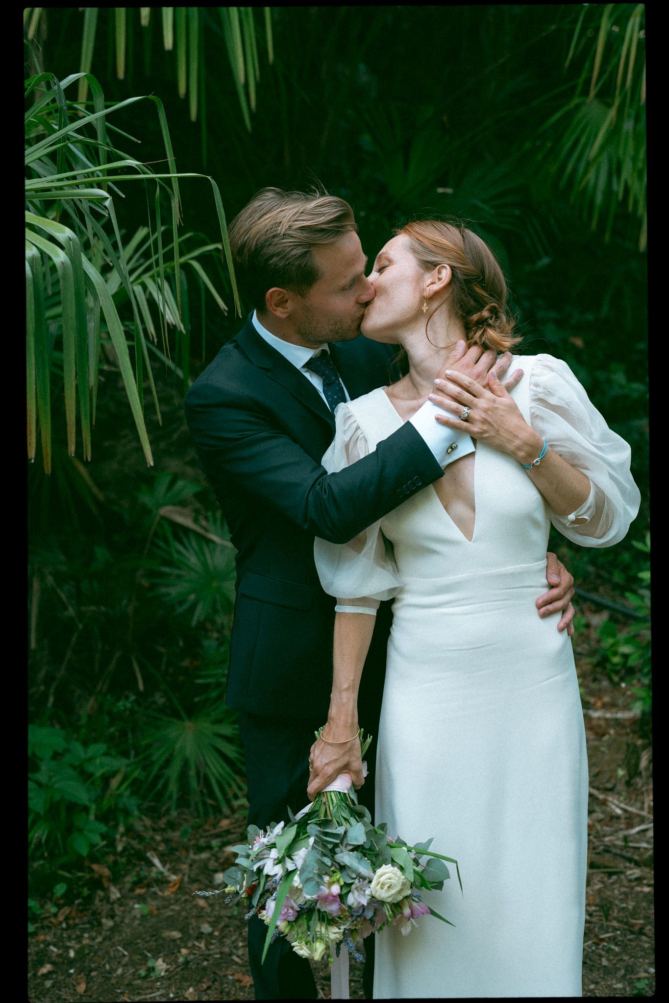 A couple in wedding attire embraces against a backdrop of tropical greenery and palm fronds.