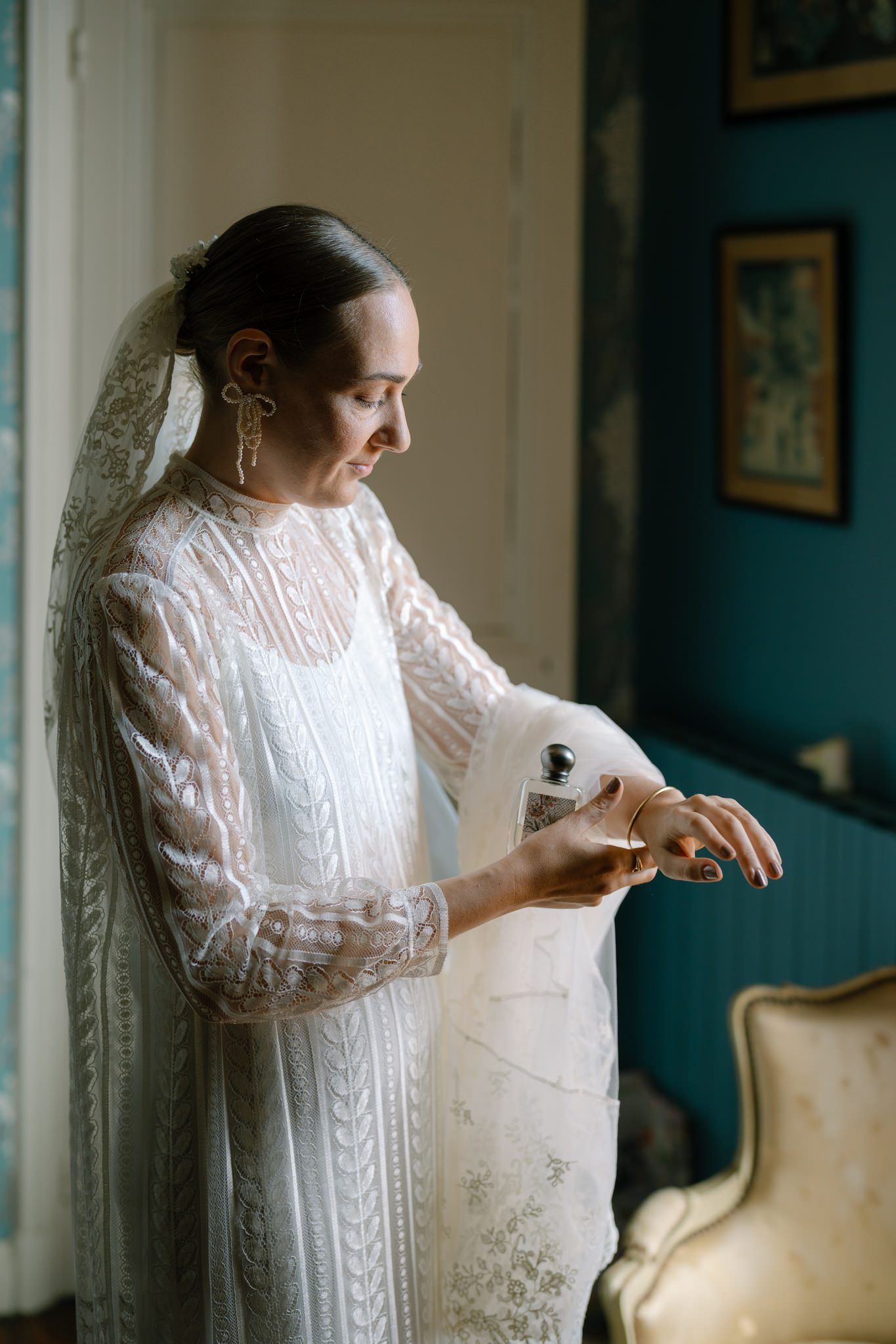Bride in long-sleeved lace wedding dress preparing in room with teal walls.