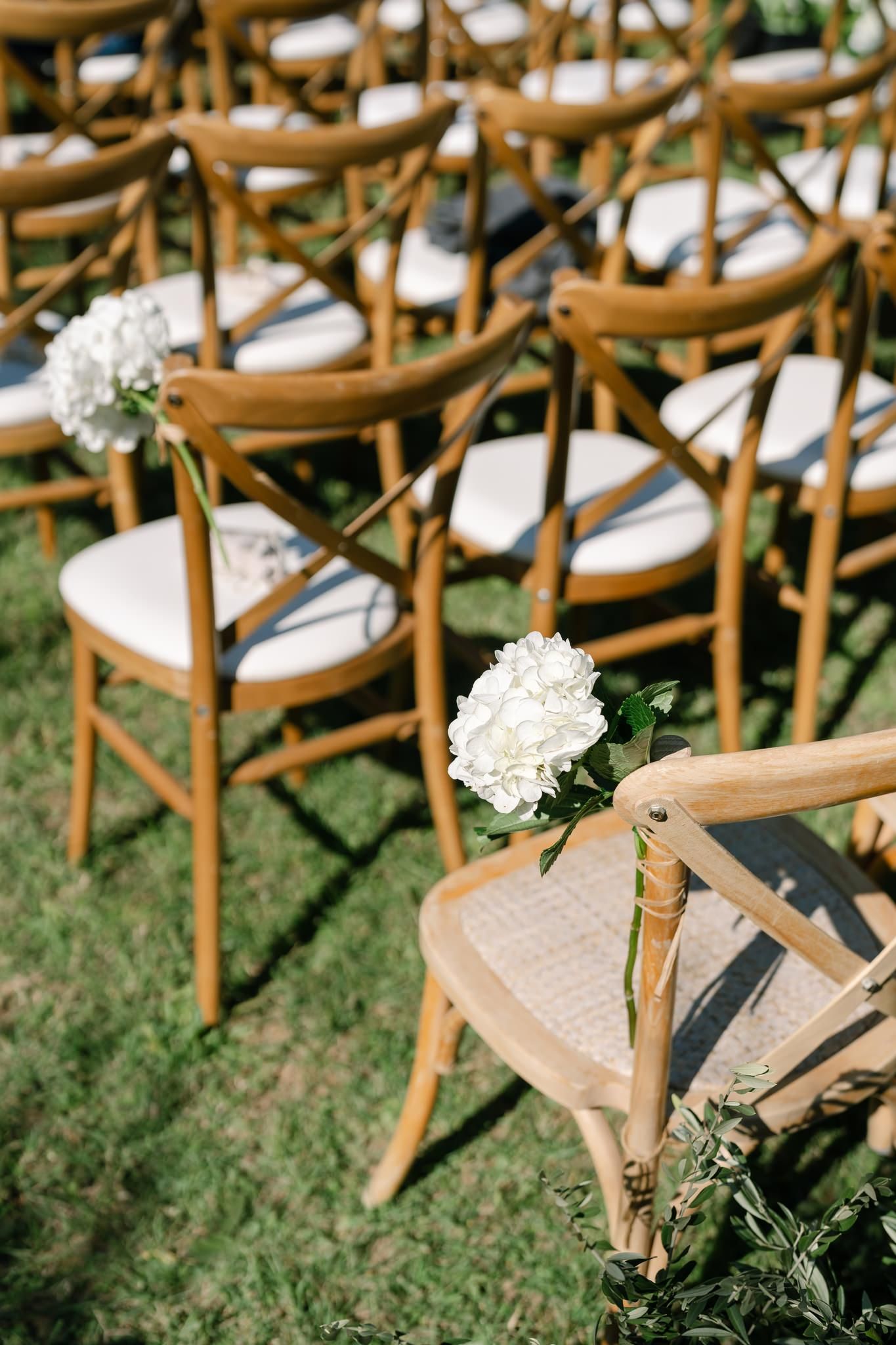Wooden cross-back chairs with white cushions and floral decorations set up for outdoor ceremony.