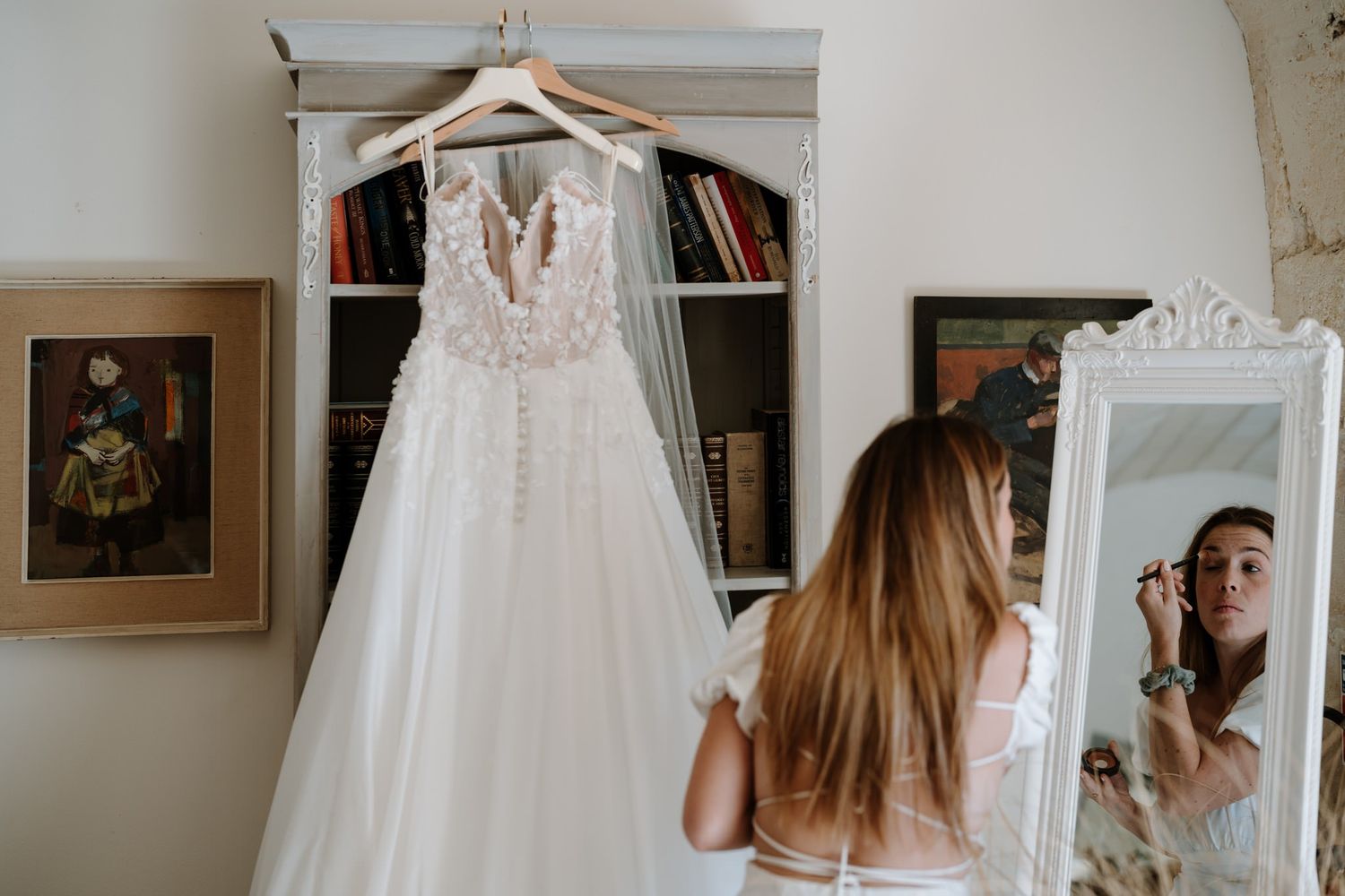 A white lace wedding dress hangs on display in front of a bookshelf and vintage mirror.