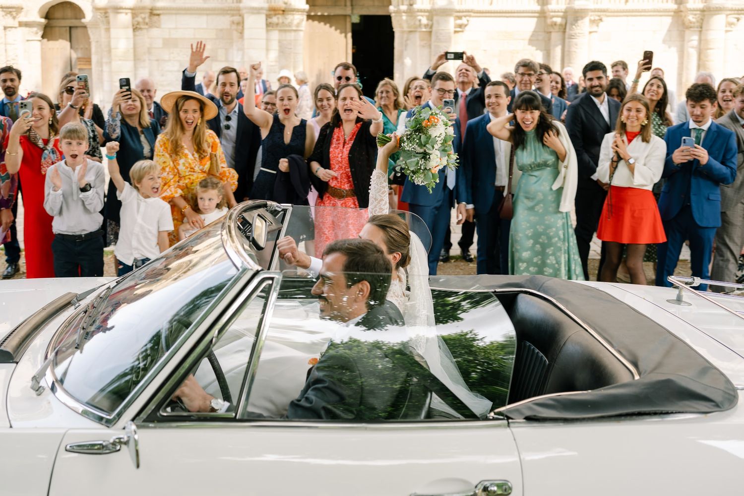 Wedding guests wave goodbye as newlyweds depart in classic car.