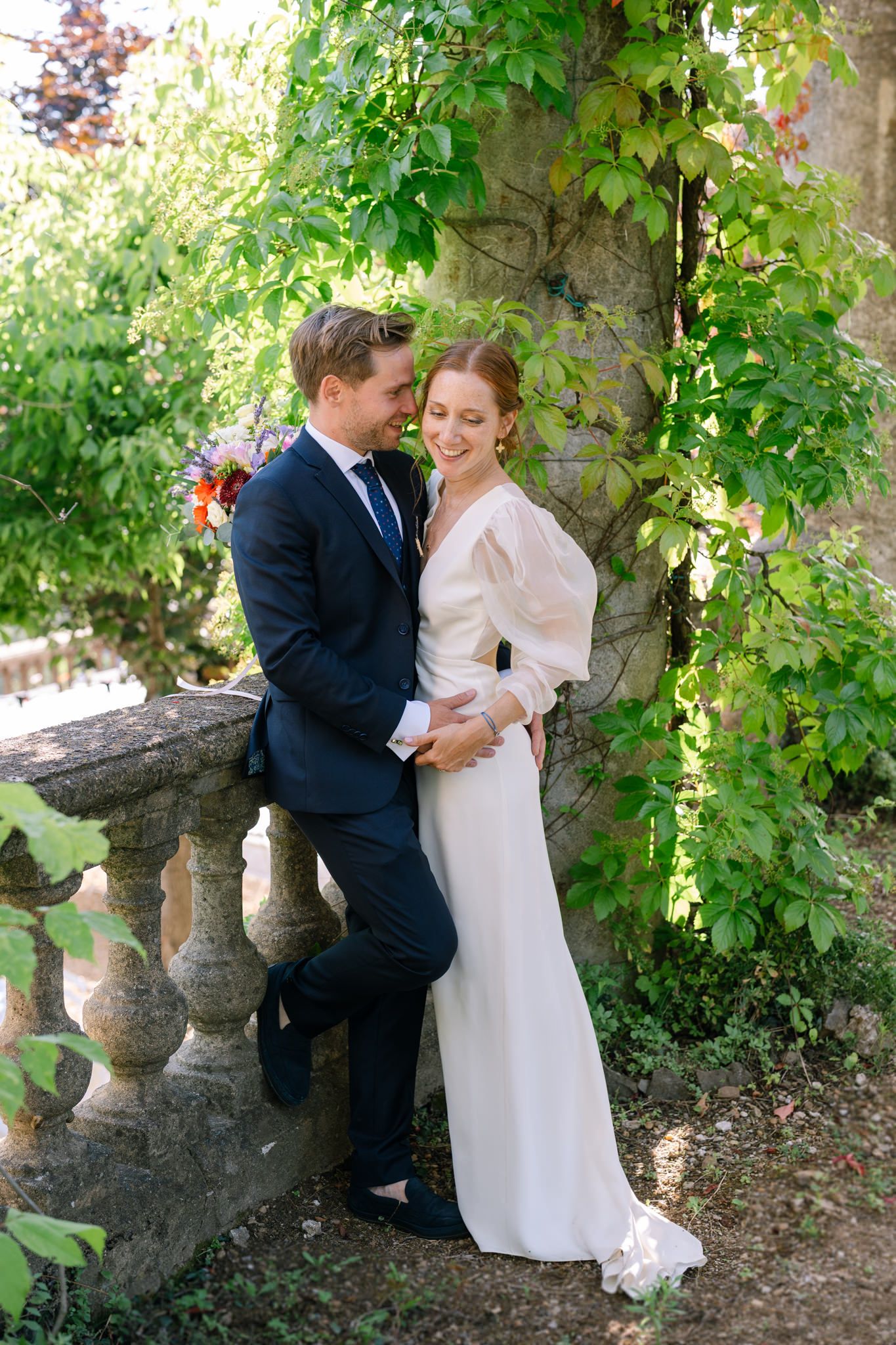 Wedding couple embraces by stone balustrade surrounded by climbing vines in garden setting.