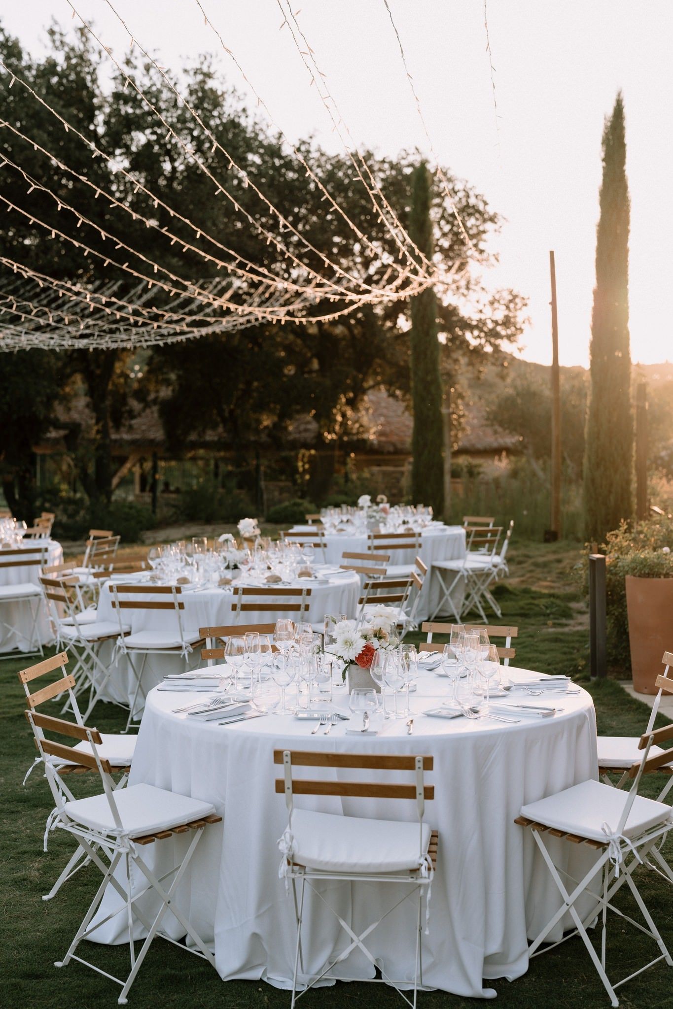 Outdoor wedding reception setup with string lights and white round tables at sunset.