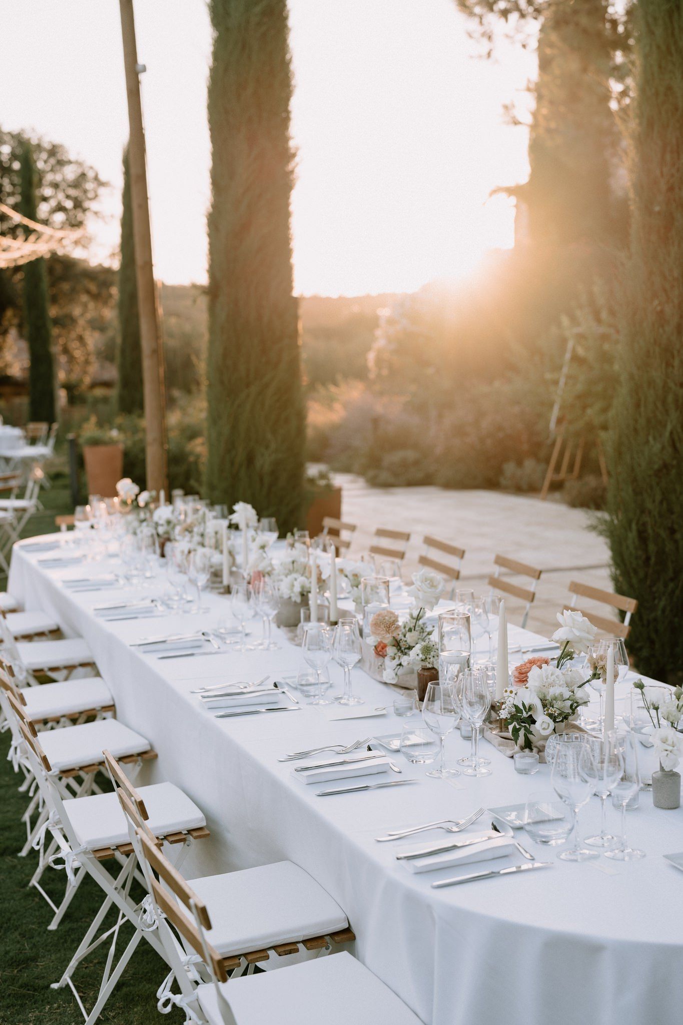 Outdoor wedding reception table set with white linens under cypress trees at sunset.