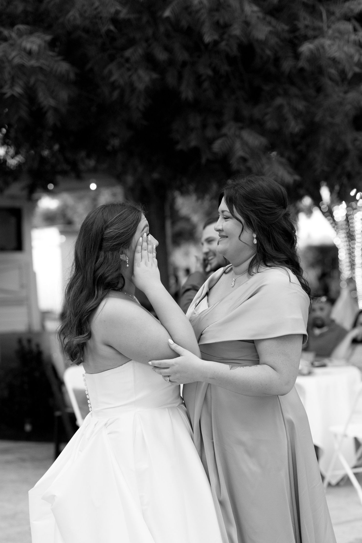 Two women share an emotional moment at a wedding, with one in a white dress and one in a gray dress outdoors.