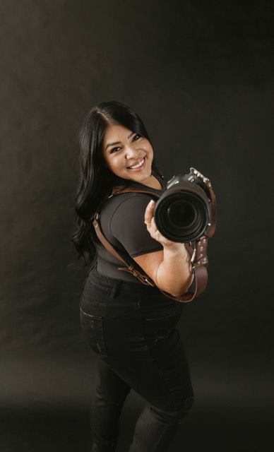 Professional photographer in black outfit holds camera while posing against dark background.