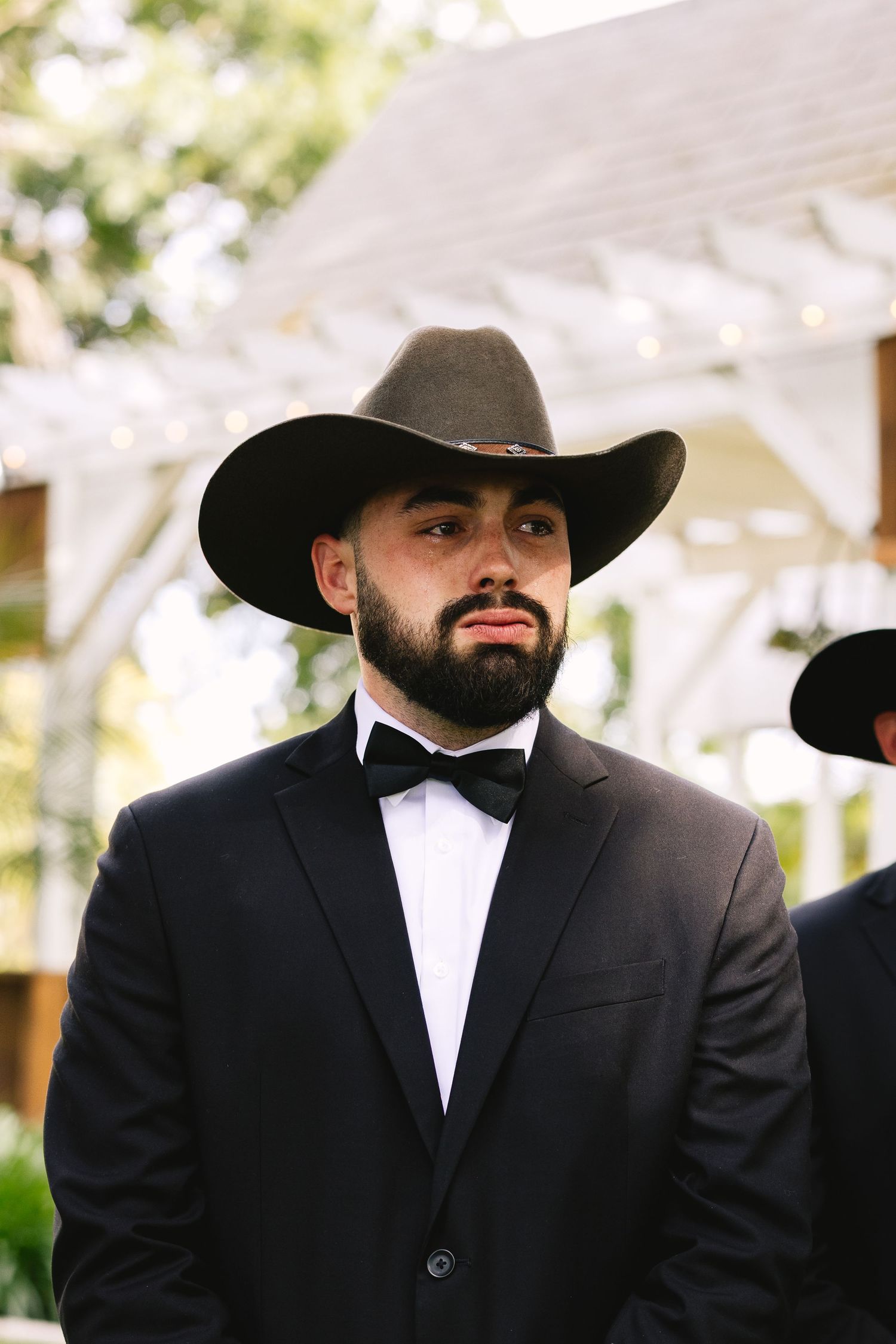 A bearded person wearing a black cowboy hat and formal tuxedo with bow tie stands outdoors at an event.