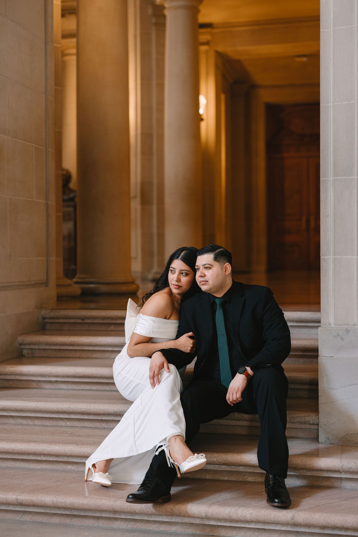 A couple in formal attire sits together on grand marble steps in an elegant hallway.
