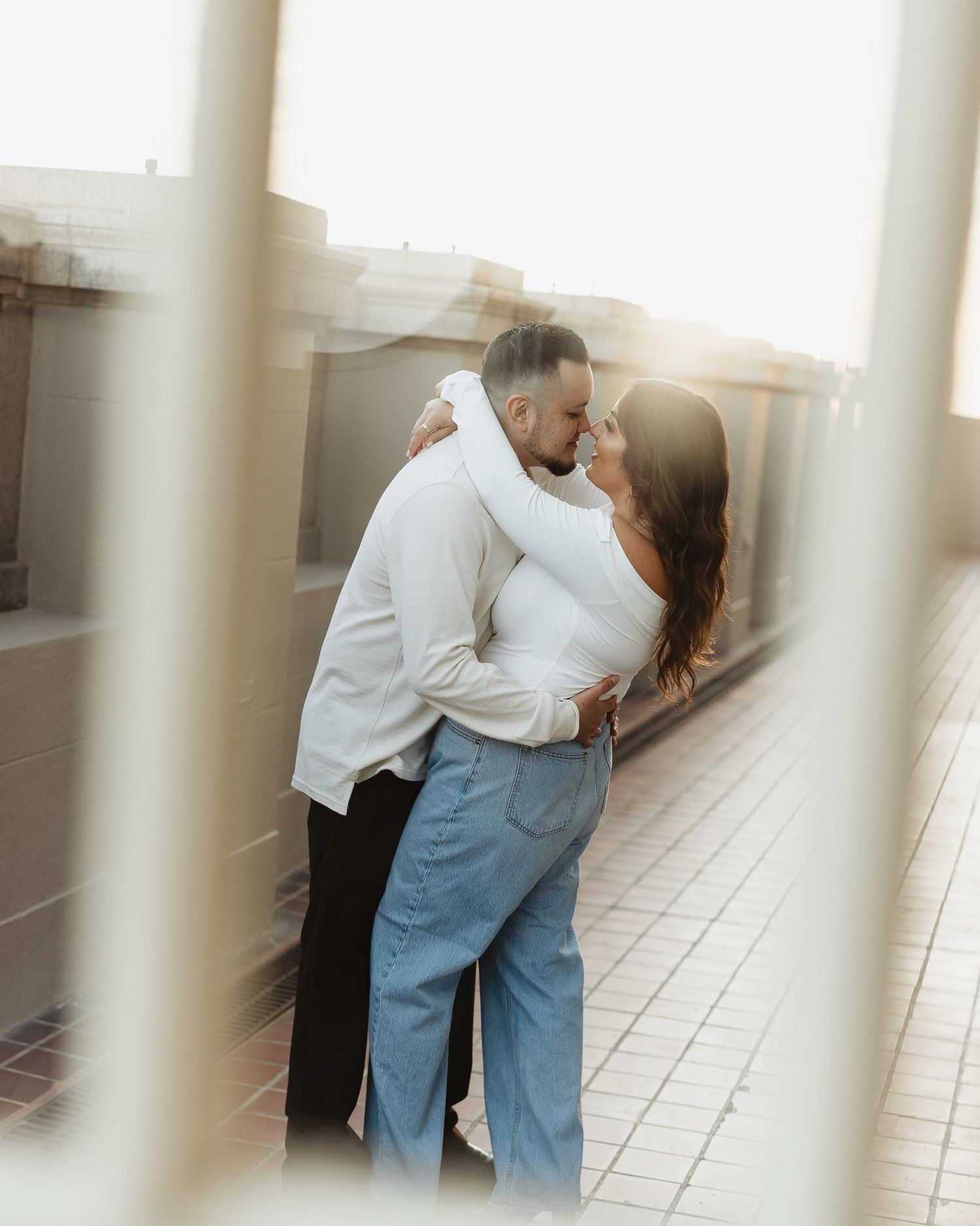 A couple shares an intimate moment while embracing on a patio, shot through window panes in soft natural light.