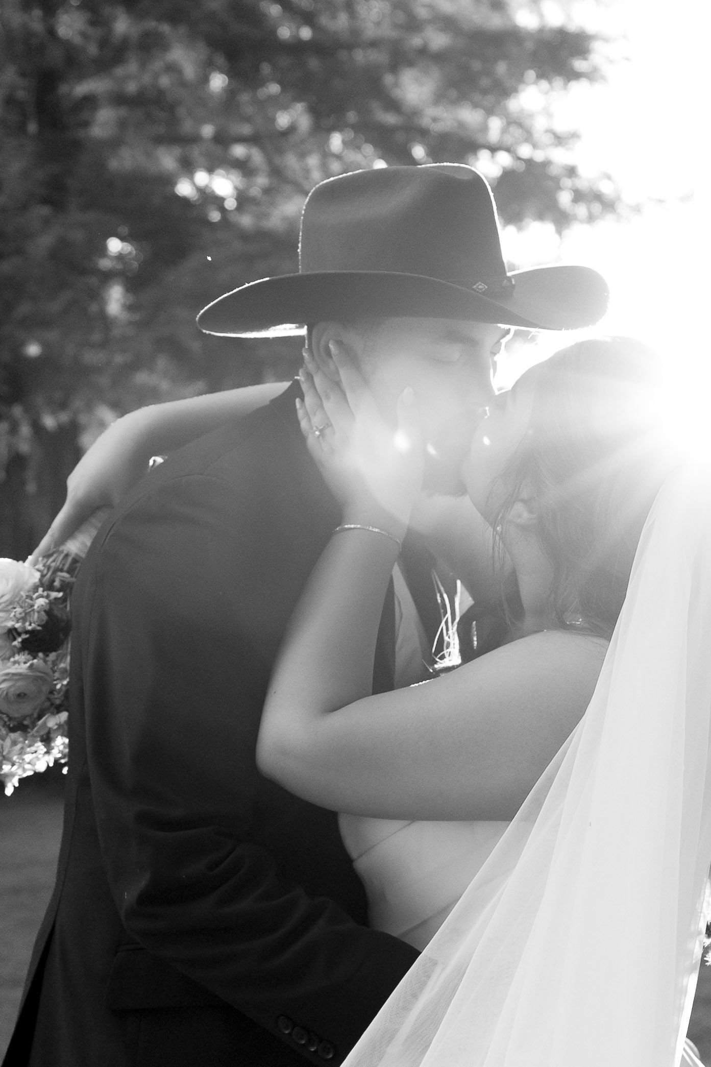 Black and white wedding photo of a couple kissing under bright sunlight with one person wearing a cowboy hat.
