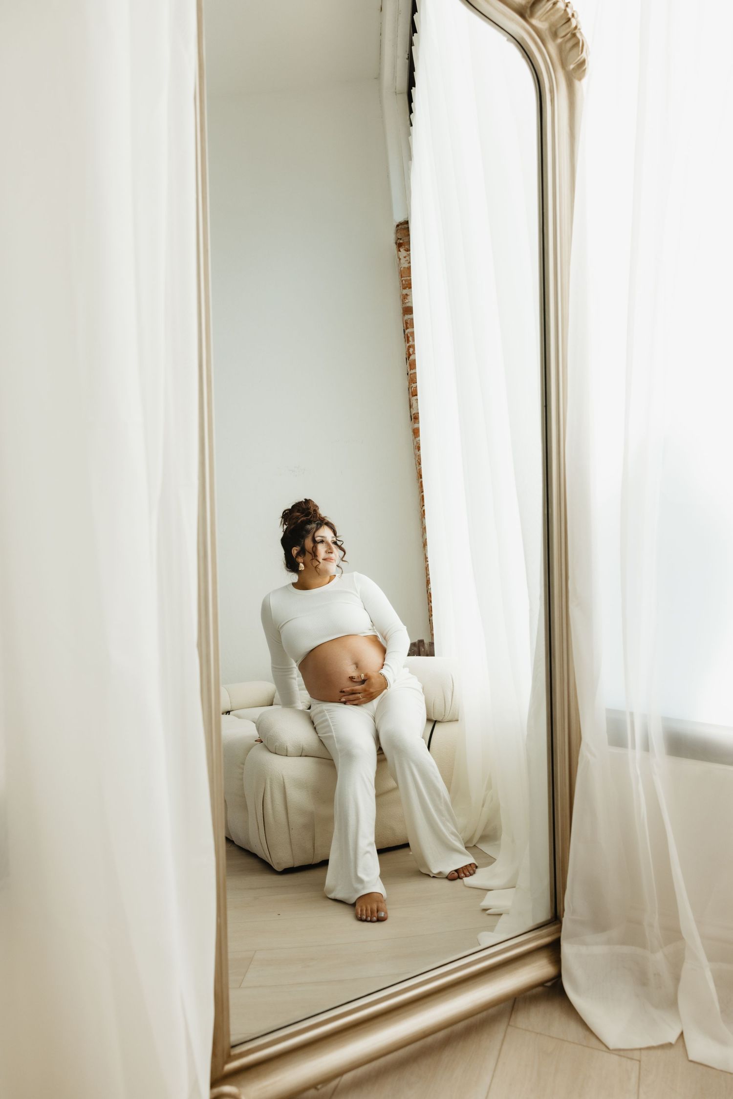 A pregnant person in white loungewear sits on a couch while being reflected in an ornate floor mirror.