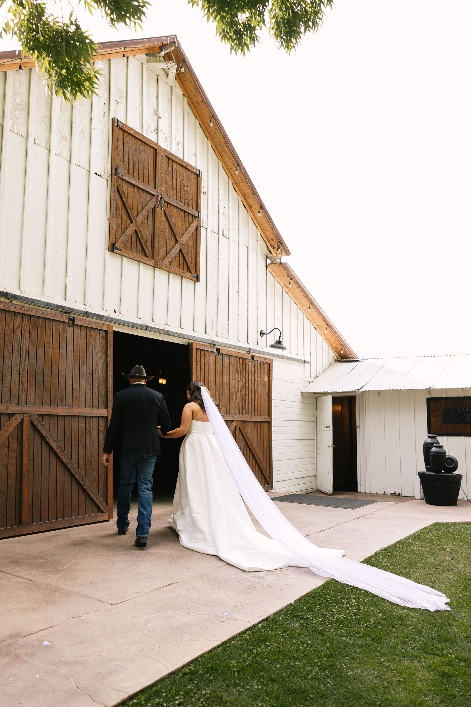 A couple enters a rustic white barn with large wooden doors while a flowing white train trails behind.