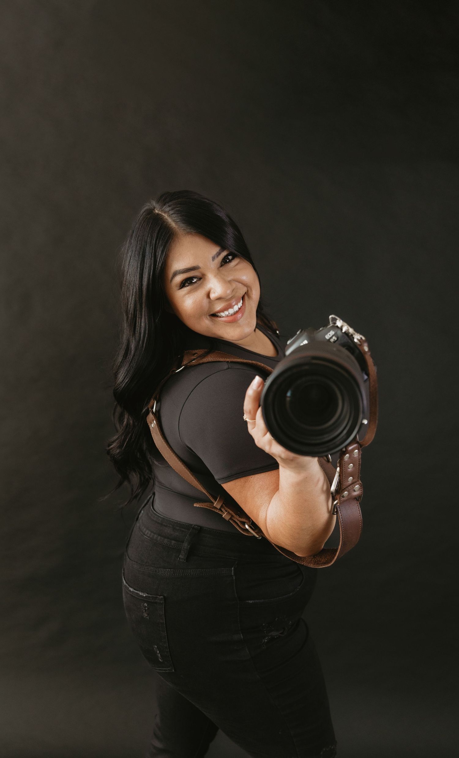Professional photographer wearing all black smiles while holding a DSLR camera against dark backdrop.
