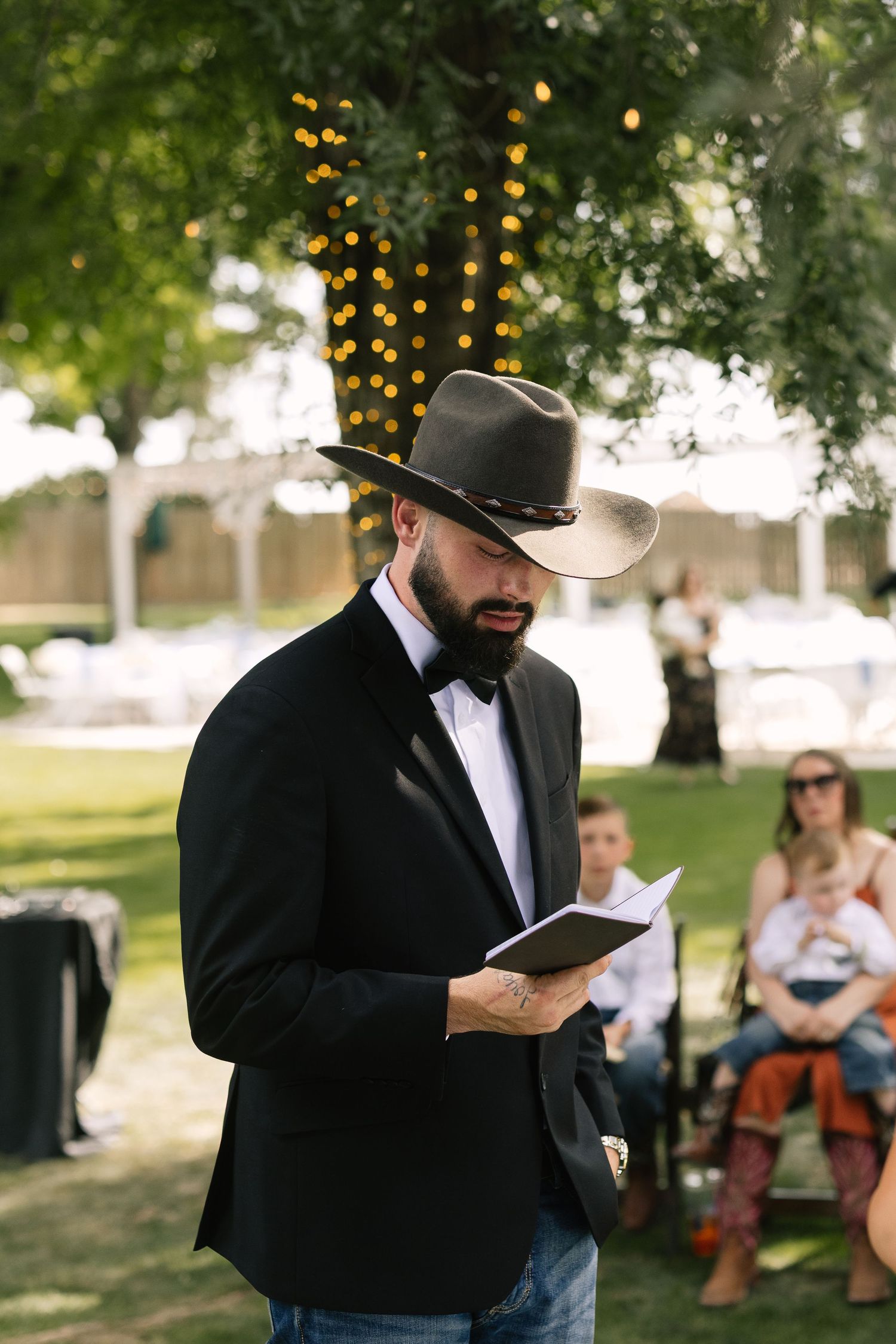 A person in black suit and hat reading from a book outdoors with string lights and trees in the background.