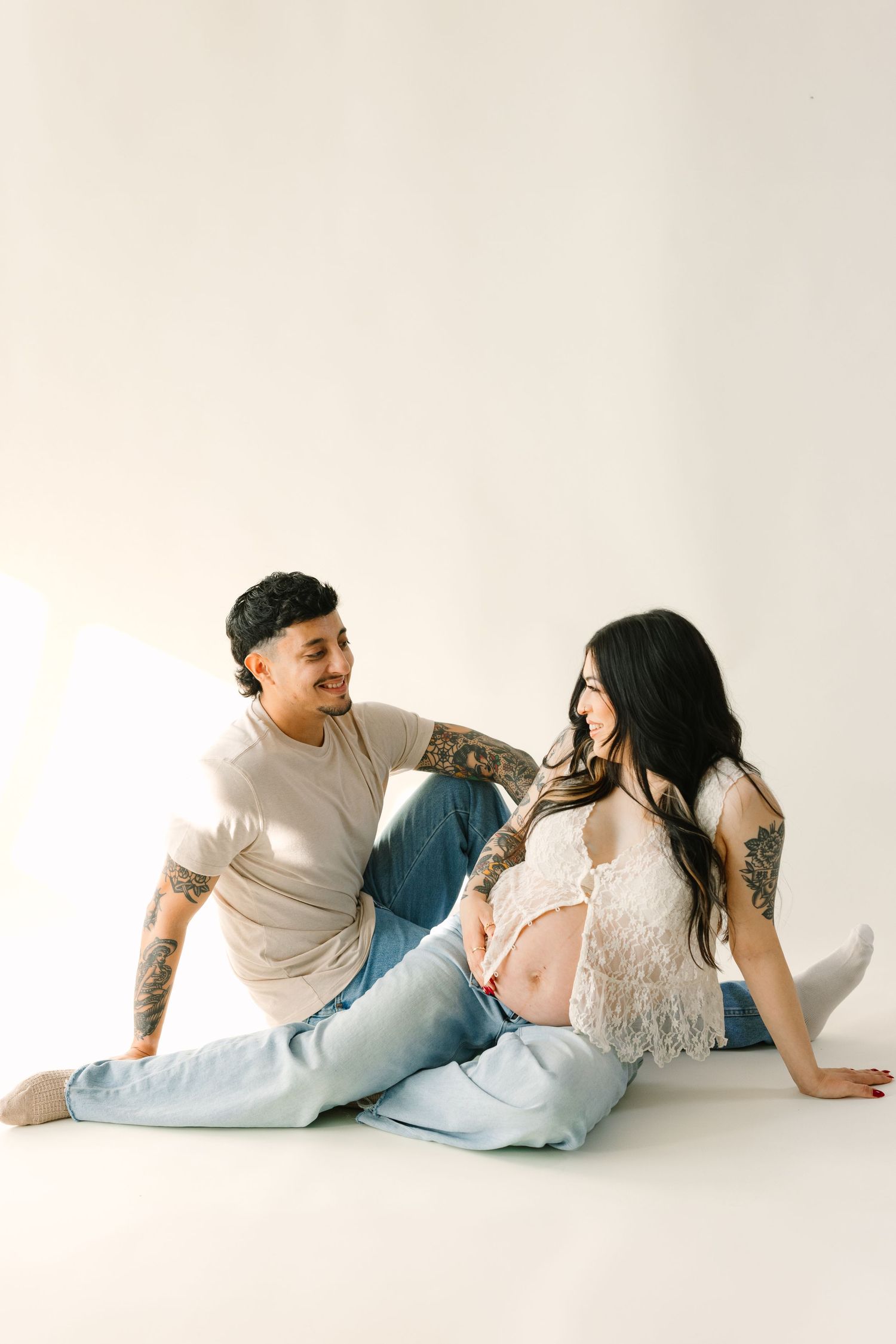 A couple in casual attire sits intimately on a white background, both wearing jeans and appearing relaxed.