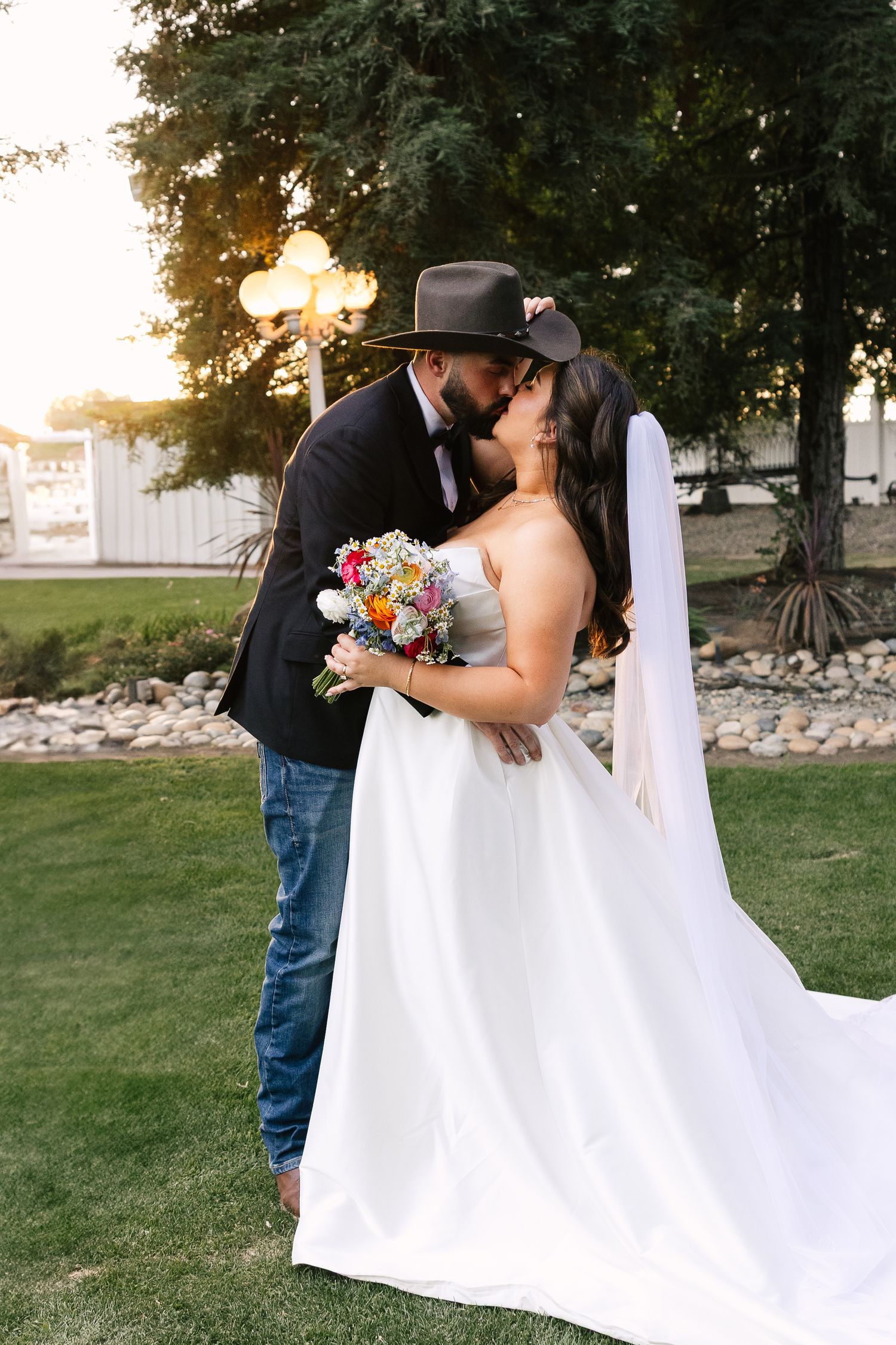 A bride in a white gown holding a colorful bouquet shares a romantic sunset moment with her cowboy-hatted groom outdoors.