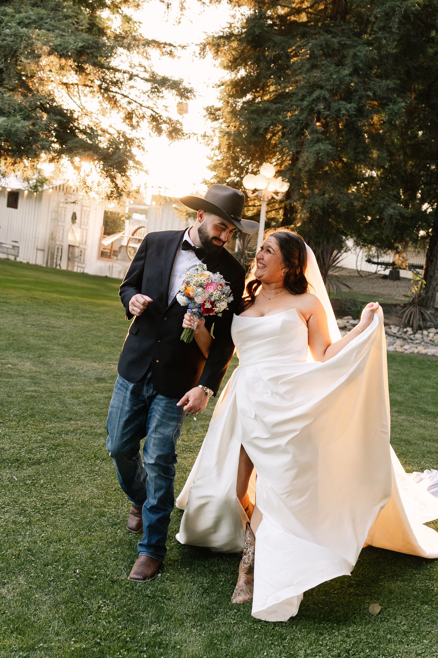 A couple celebrates their wedding day outdoors at sunset, with the bride in white and the groom in black and denim.