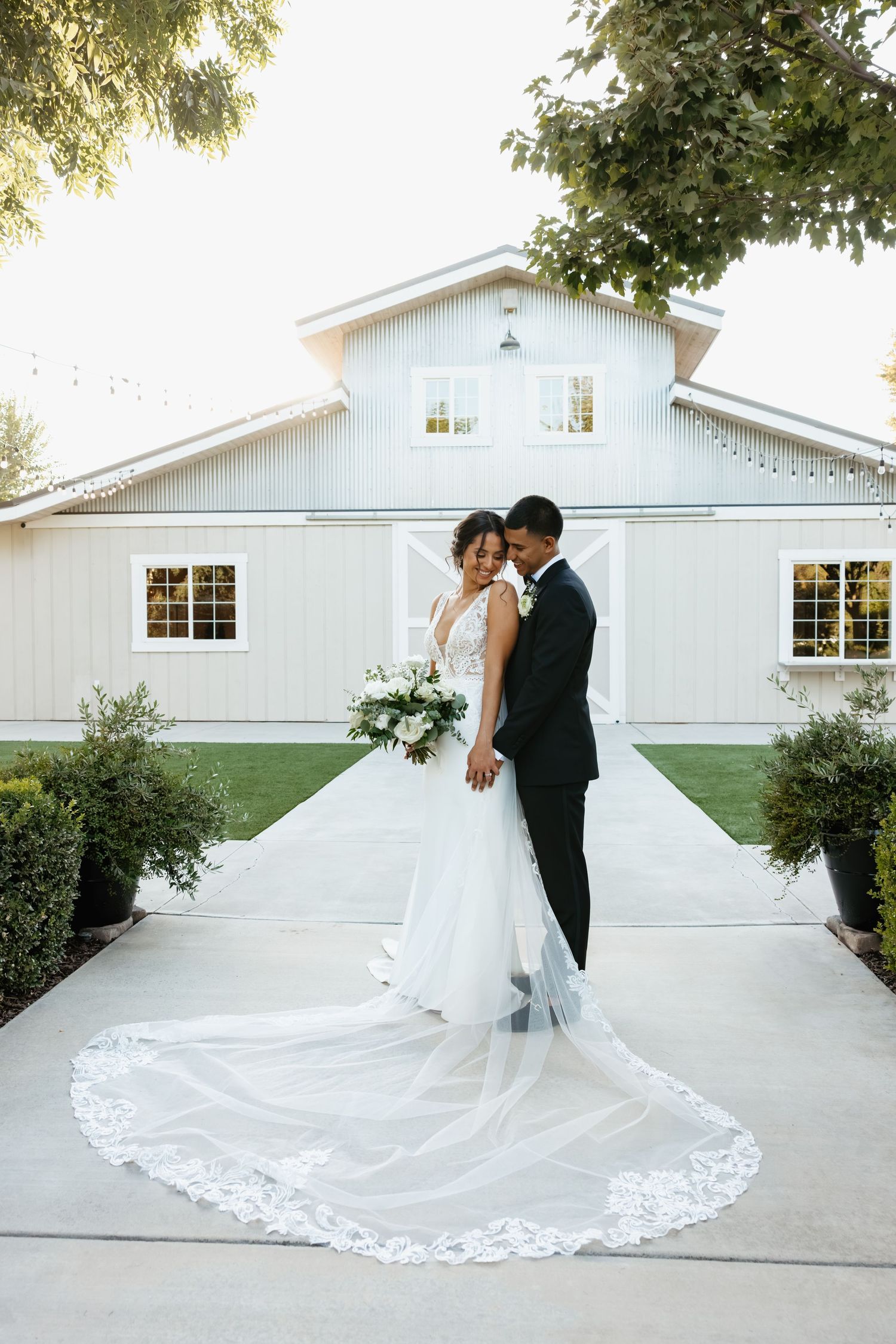 Elegant wedding portrait in front of a white barn with dramatic cathedral veil detail.