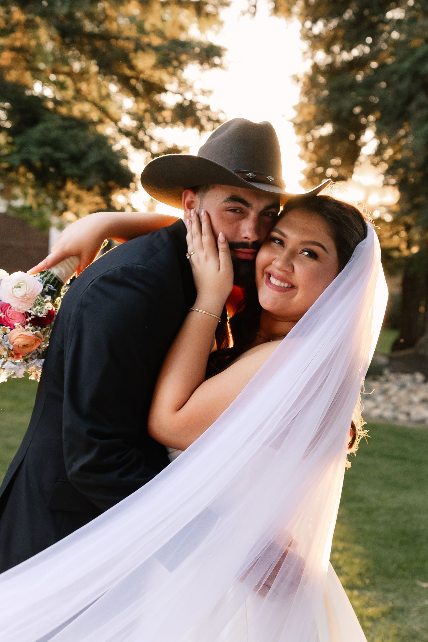 A romantic wedding couple embraces outdoors at sunset with sunlight filtering through trees and flowing white veil.