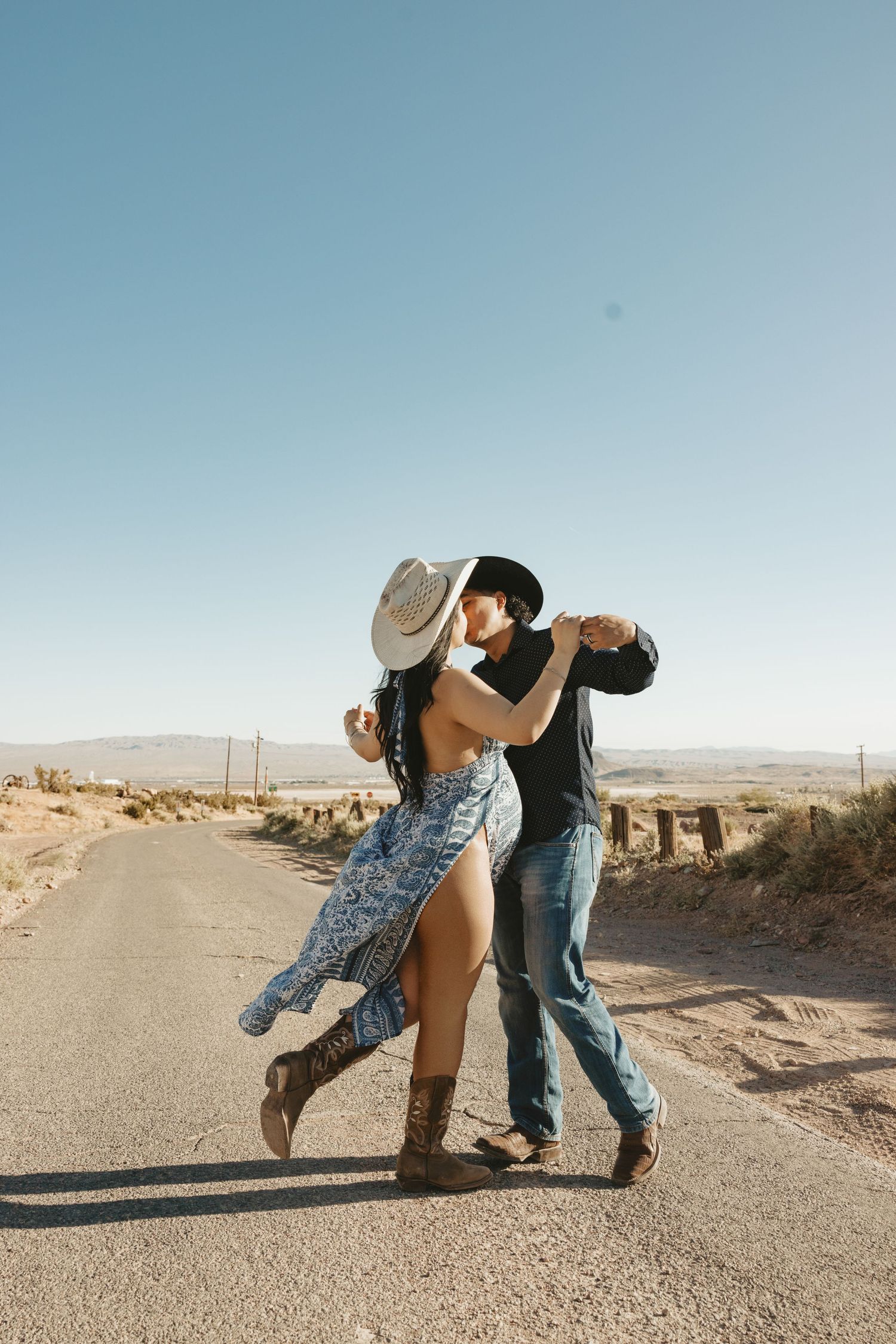 Couple dancing romantically on a desert road with cowboy hats and western attire.