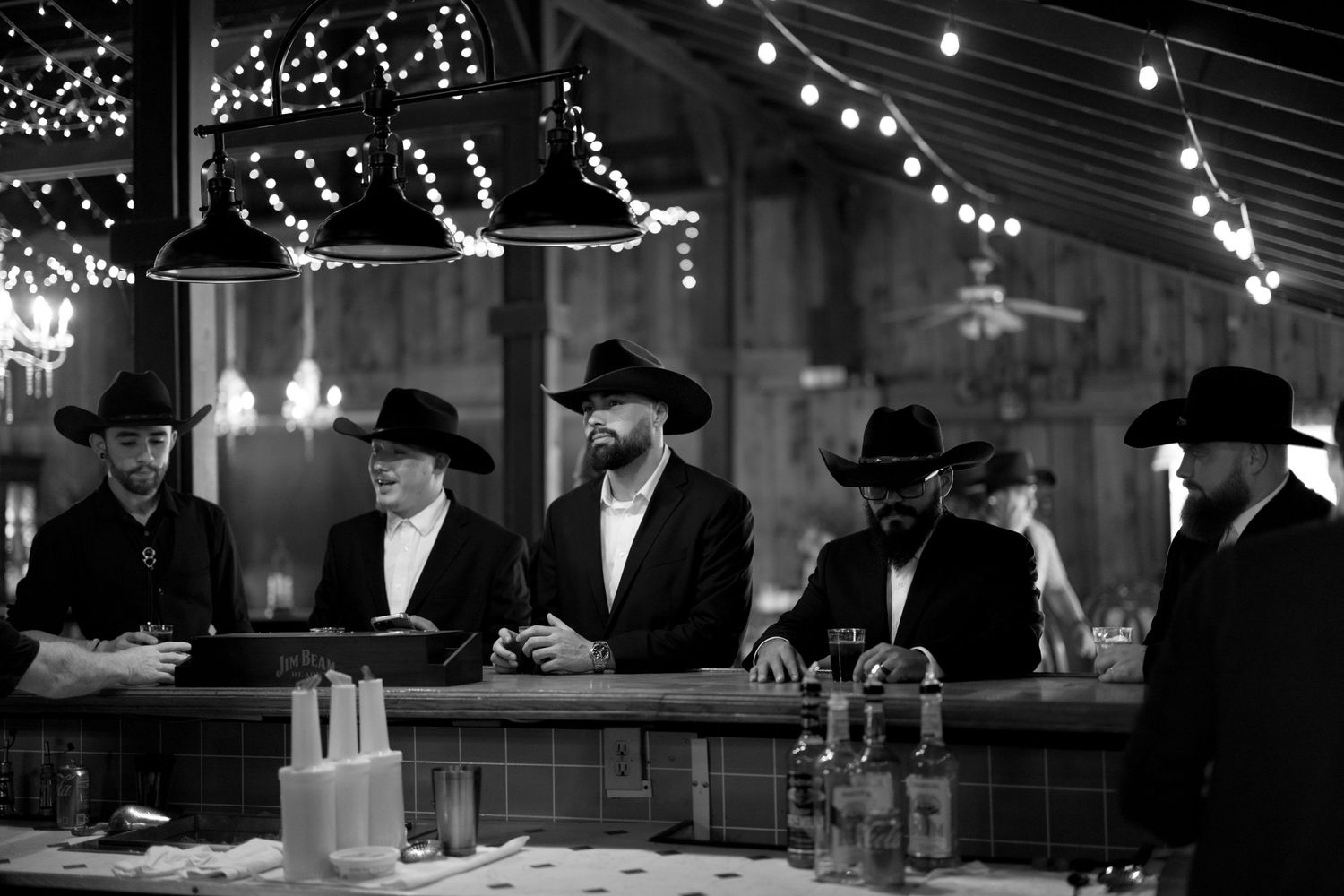 Five people in black hats and formal attire gather at a dimly lit bar adorned with string lights overhead.