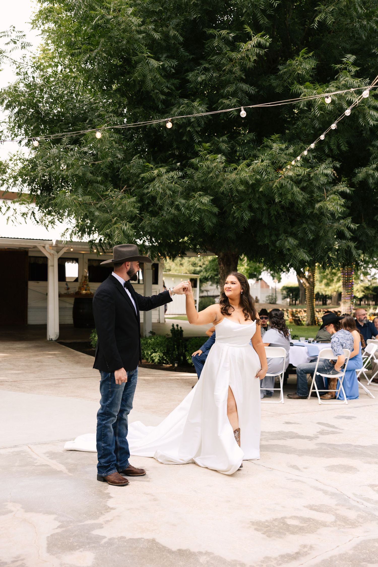 Couple dancing at outdoor wedding reception under string lights and trees on paved patio area.