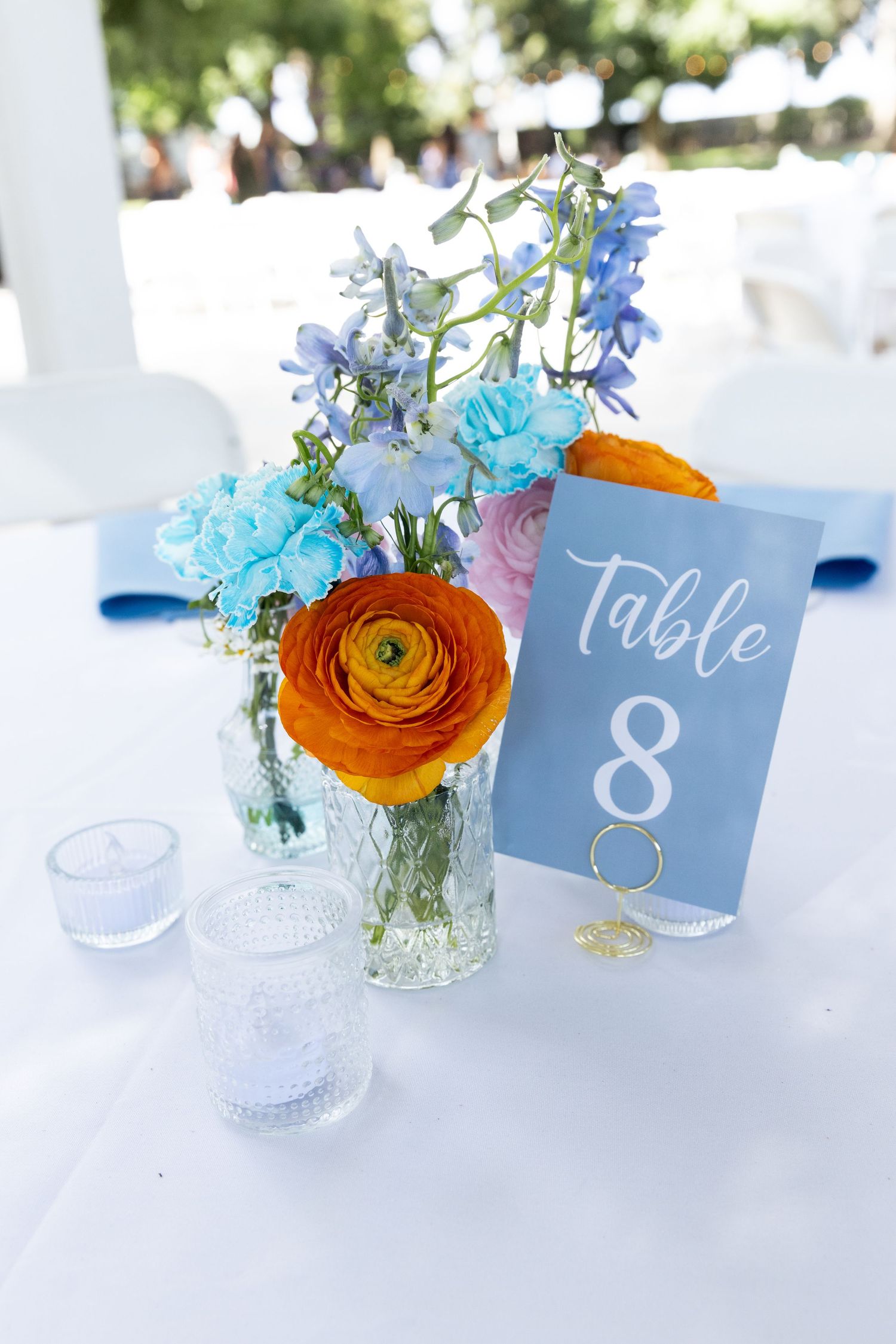 Table centerpiece with vibrant orange ranunculus and blue flowers in a glass vase next to a blue table number card.